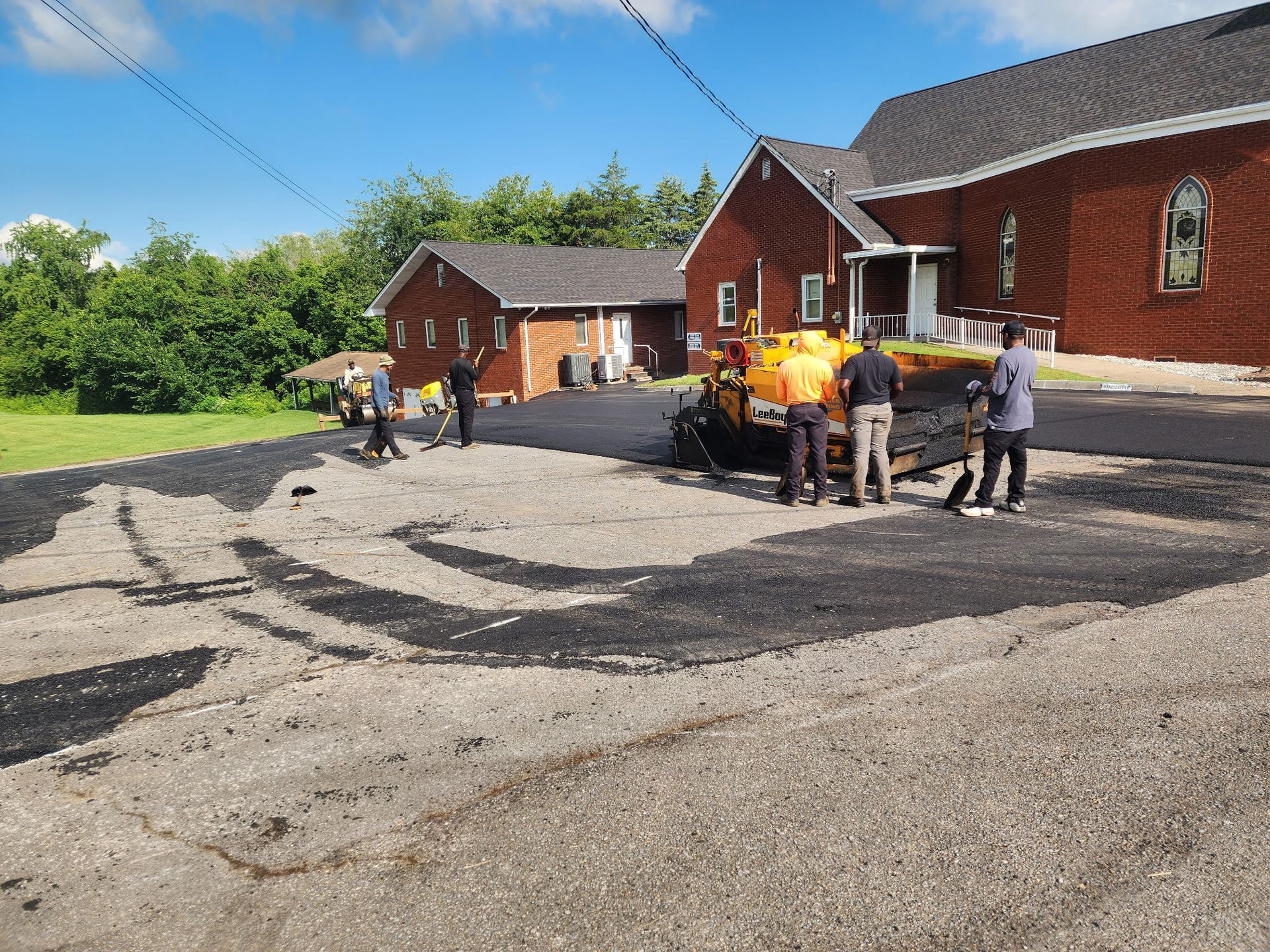 Paving crew working on asphalt near a brick church under a blue sky.