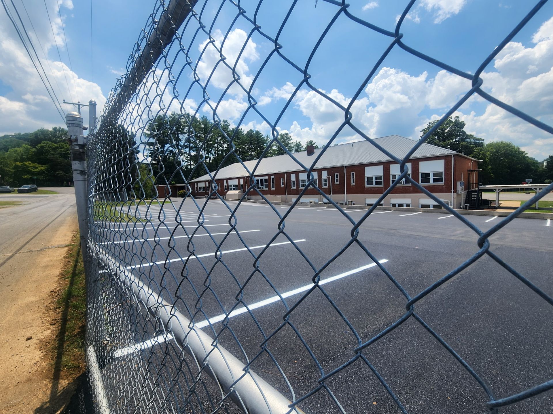 Chain-link fence in foreground, parking lot and brick building with white trim and windows in the background.