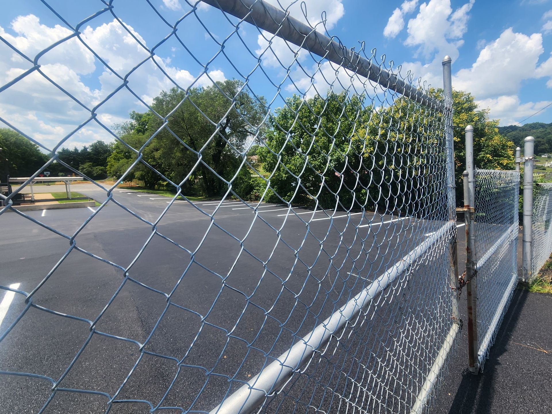 Chain-link fence in focus, overlooking a mostly empty parking lot and trees under a bright blue sky.