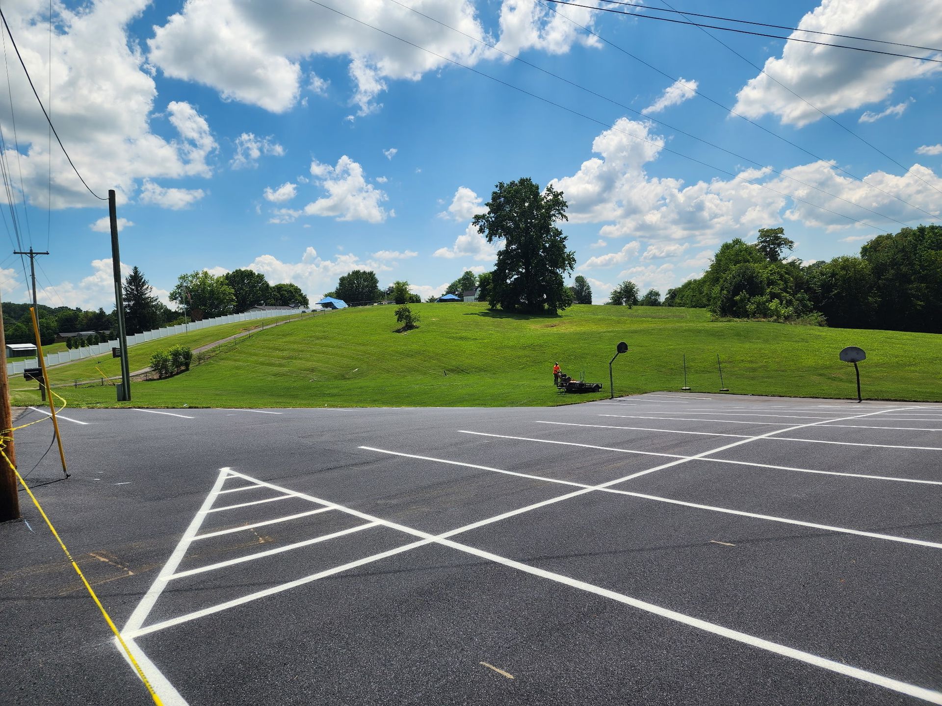 Paved parking area with white lines, leading towards a grassy hill and trees under a blue sky with clouds.