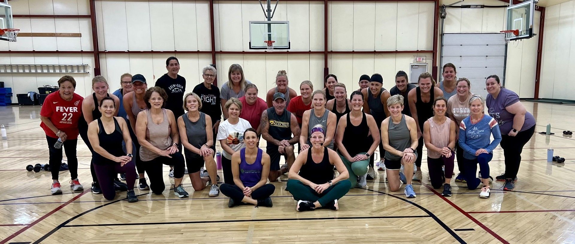 Group of people in fitness attire posing in a gym, with some kneeling.