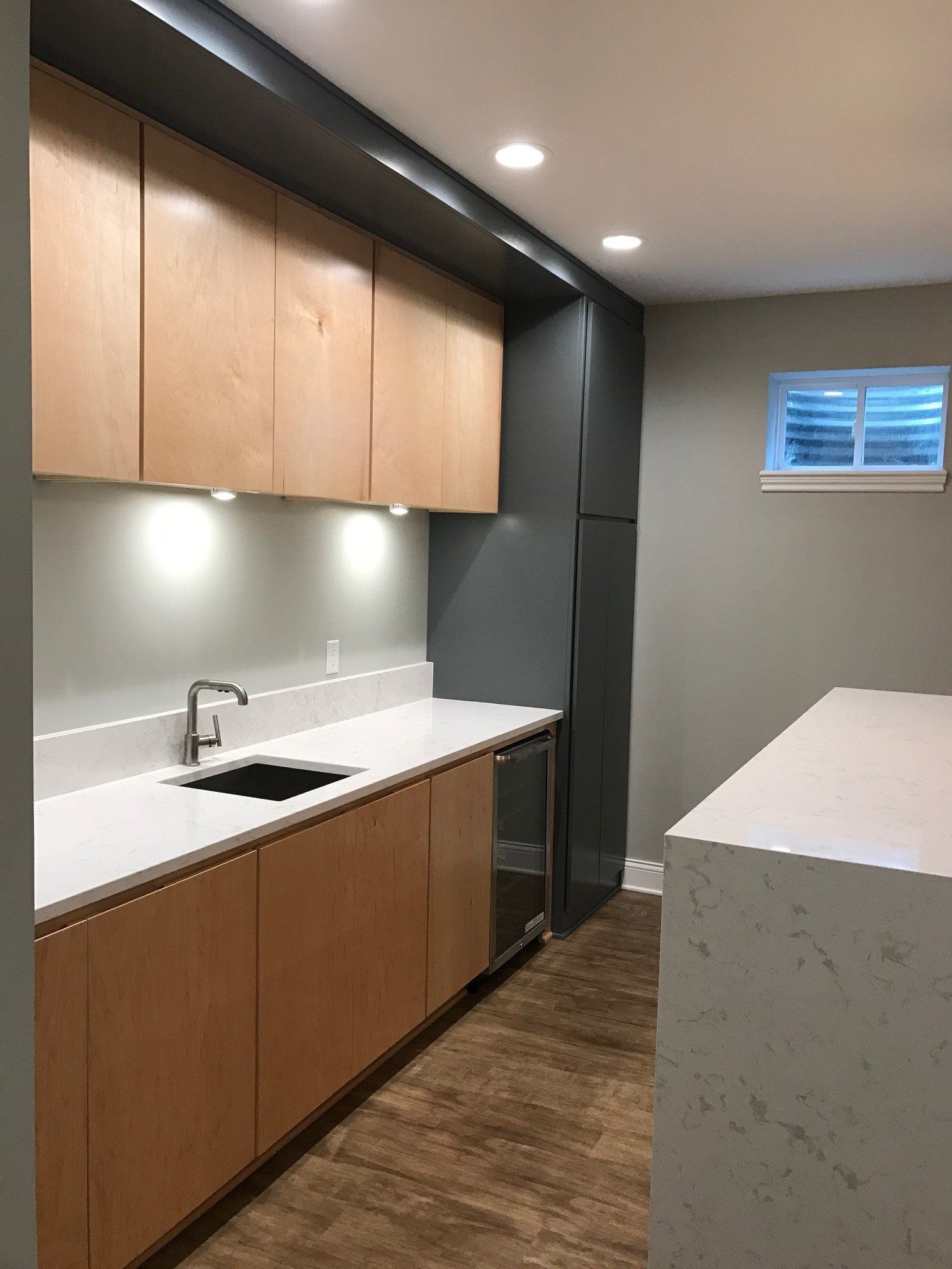 A basement kitchen with wooden cabinets , white counter tops and a sink.