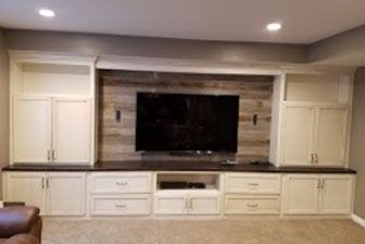 Built-in entertainment center with a TV, cabinets, drawers, and wood-paneled accent wall; cream and brown tones.
