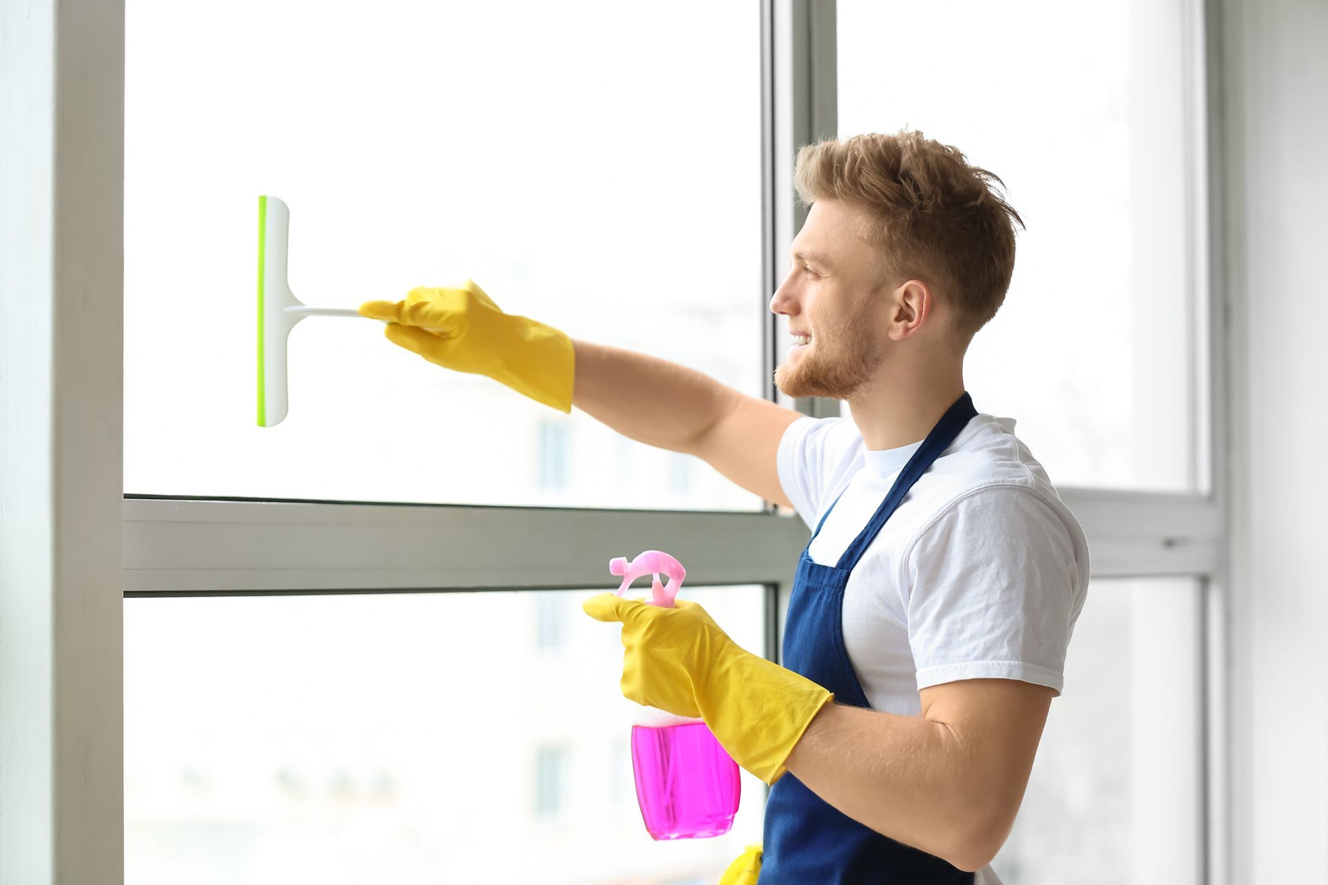 Man in gloves cleaning a window with a squeegee and pink spray bottle