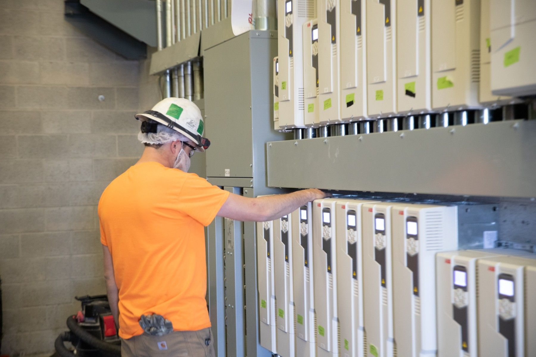 A man in an orange shirt and hard hat is working on a electrical box.