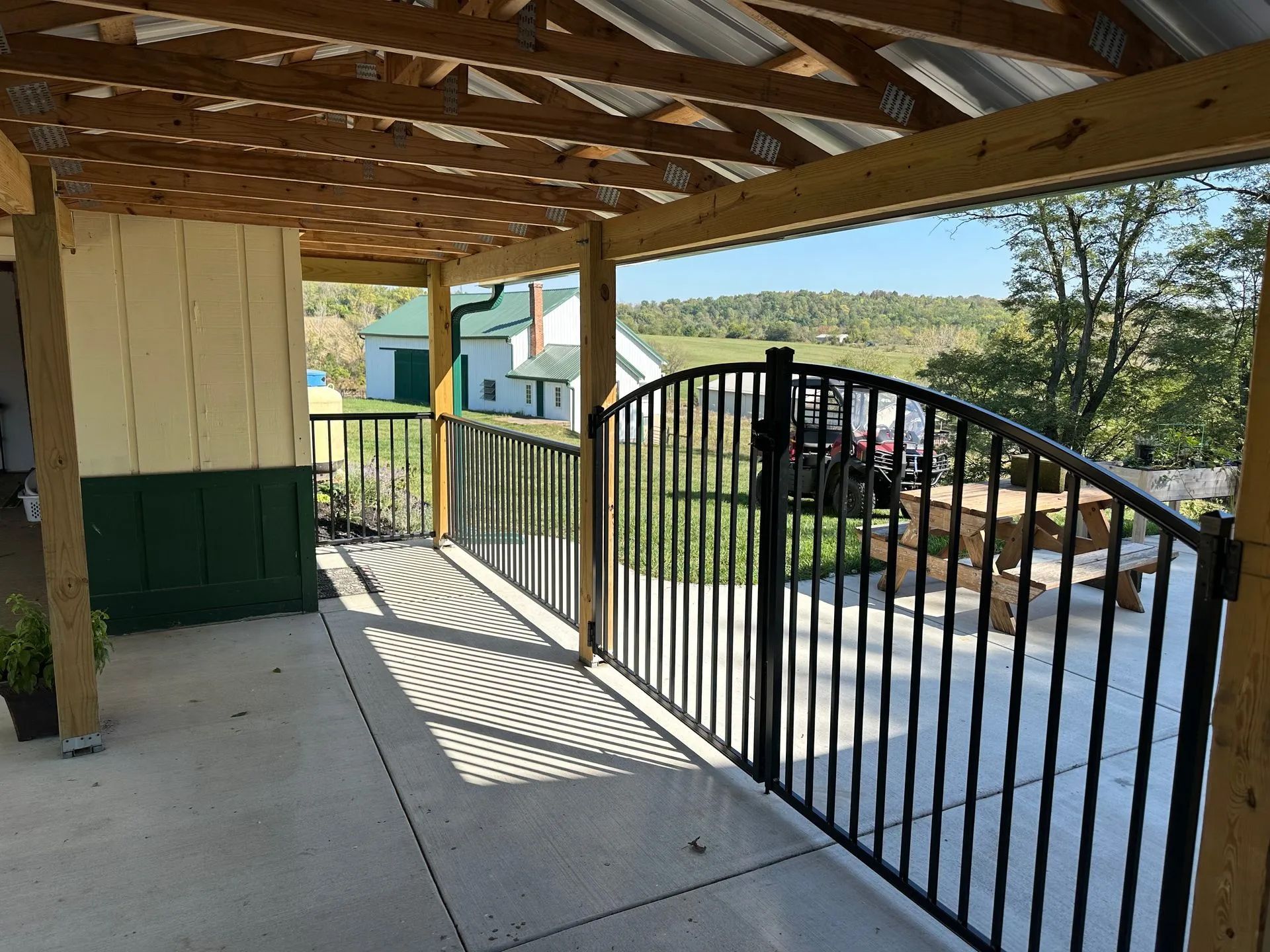 Covered patio with black metal fence, overlooking a grassy area and hills under a blue sky.