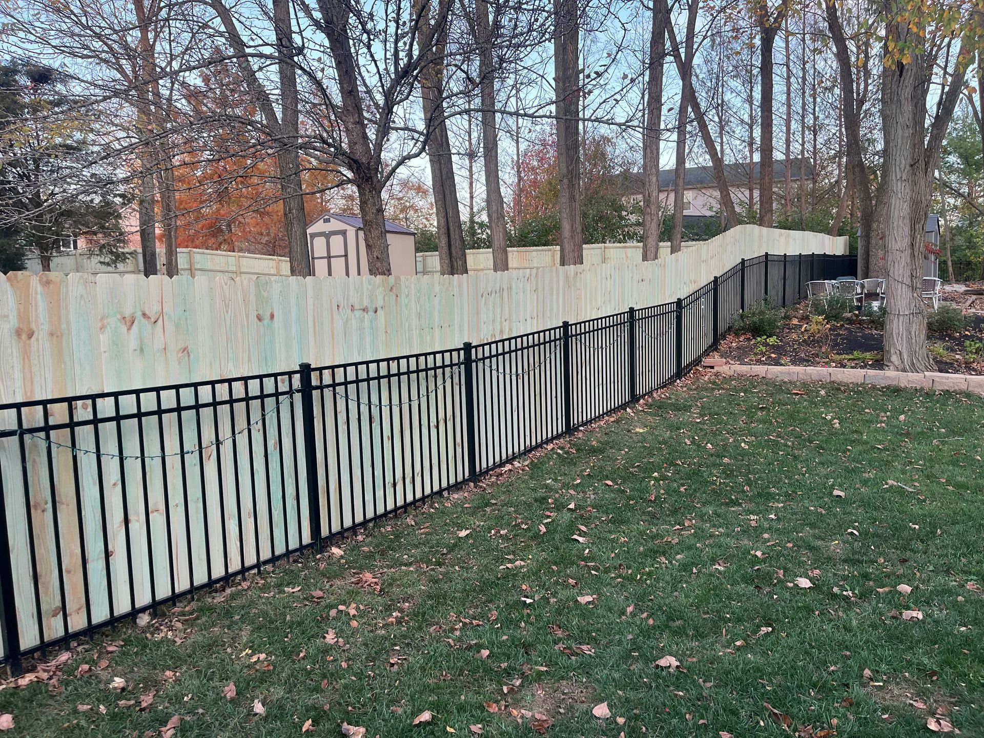 A split fence, black metal and wood, encloses a backyard lawn, trees in the background.