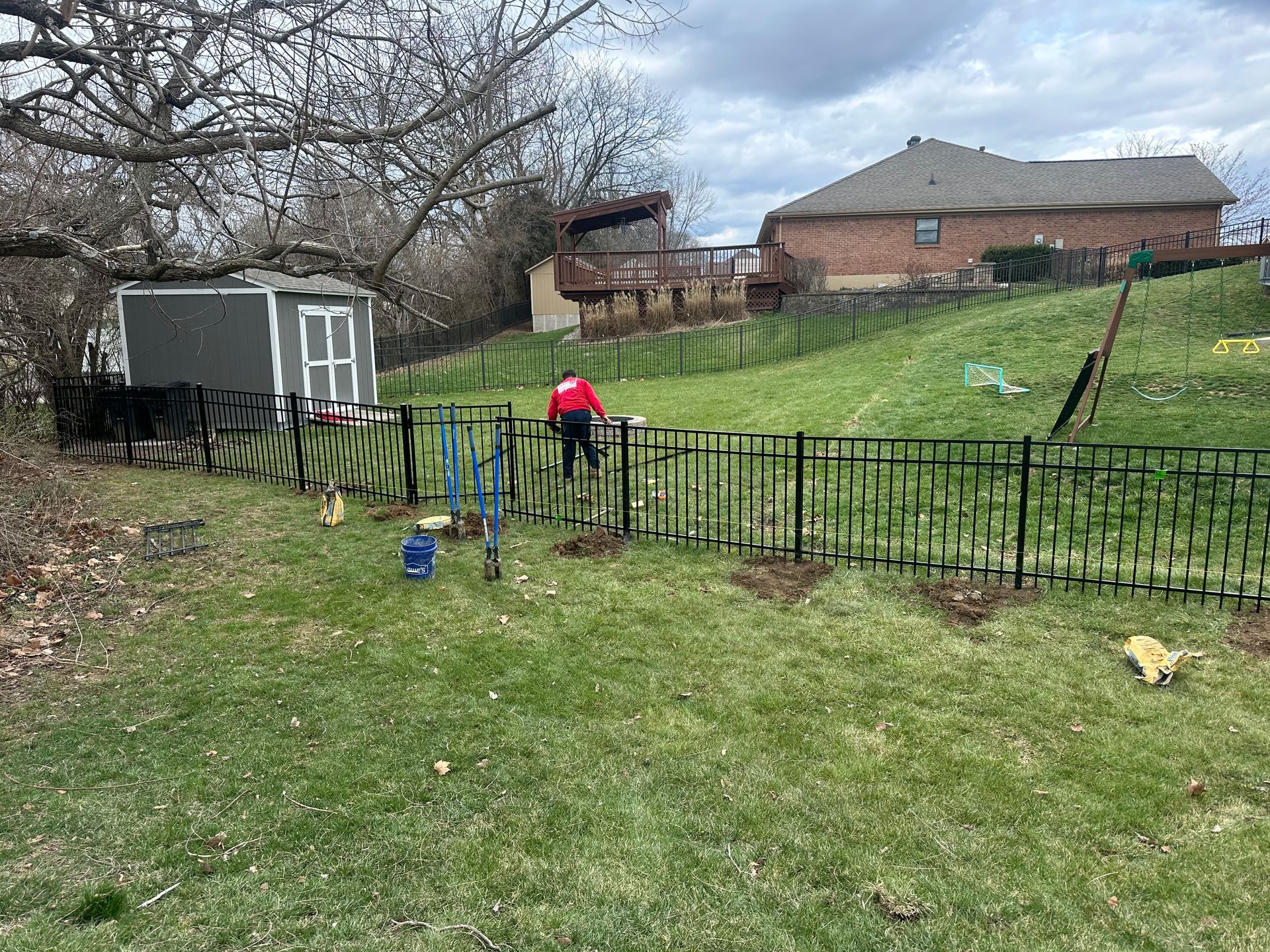 A person installing a black metal fence in a grassy backyard. A shed and house are in the background.