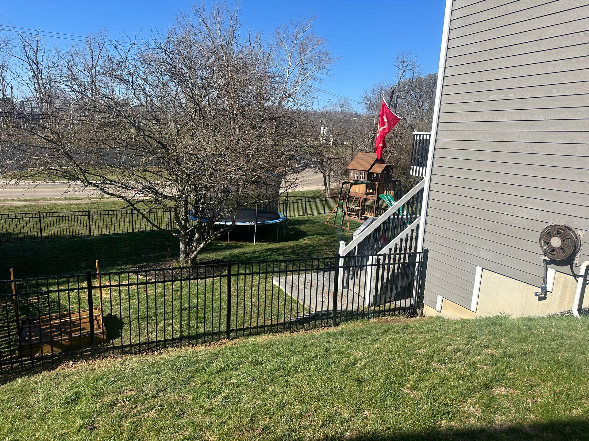 Lawn with black fence, leafless tree, and a playground visible behind a house on a sunny day.
