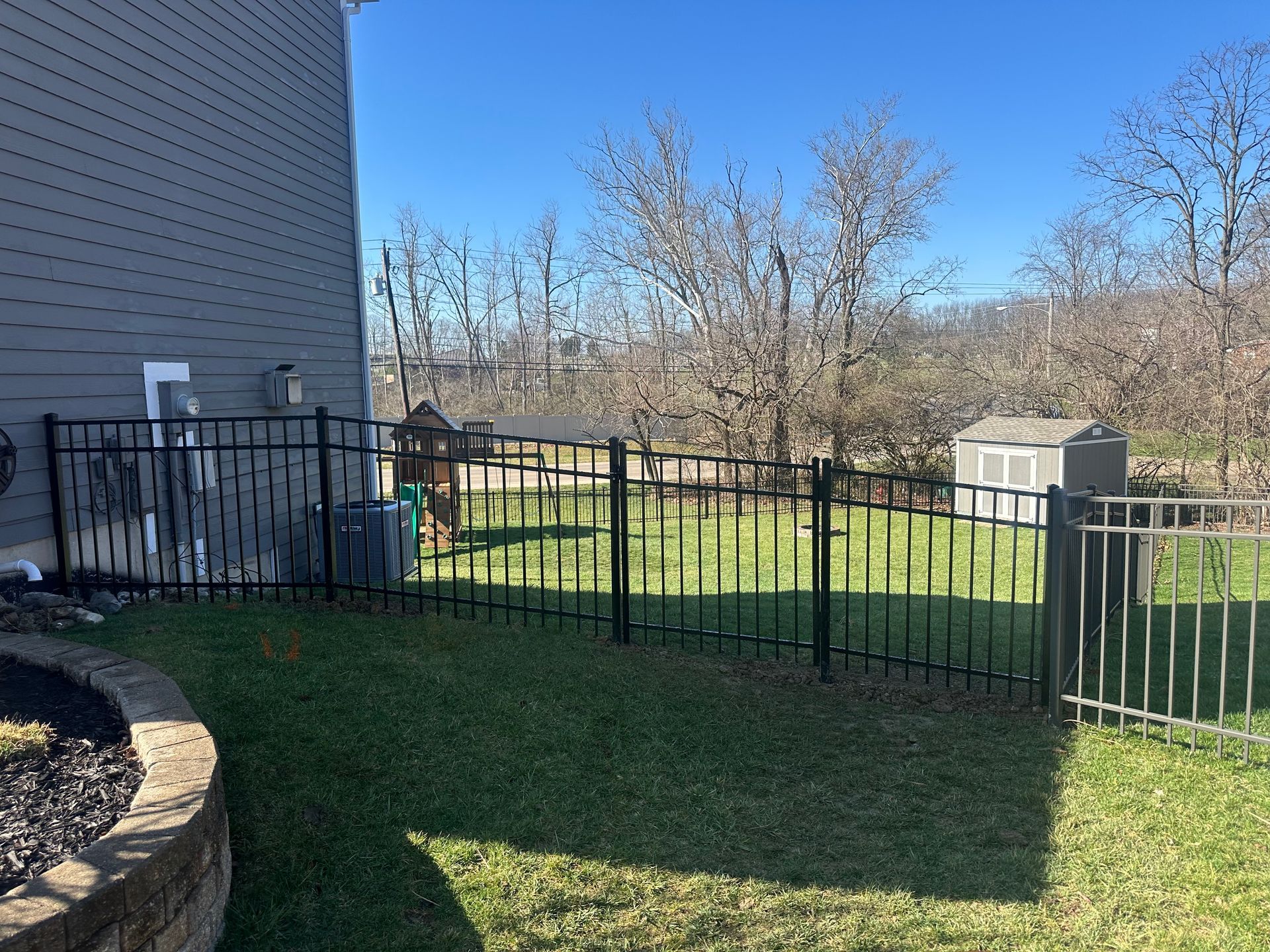 Black metal fence encloses a backyard with green grass and a small shed on a sunny day.