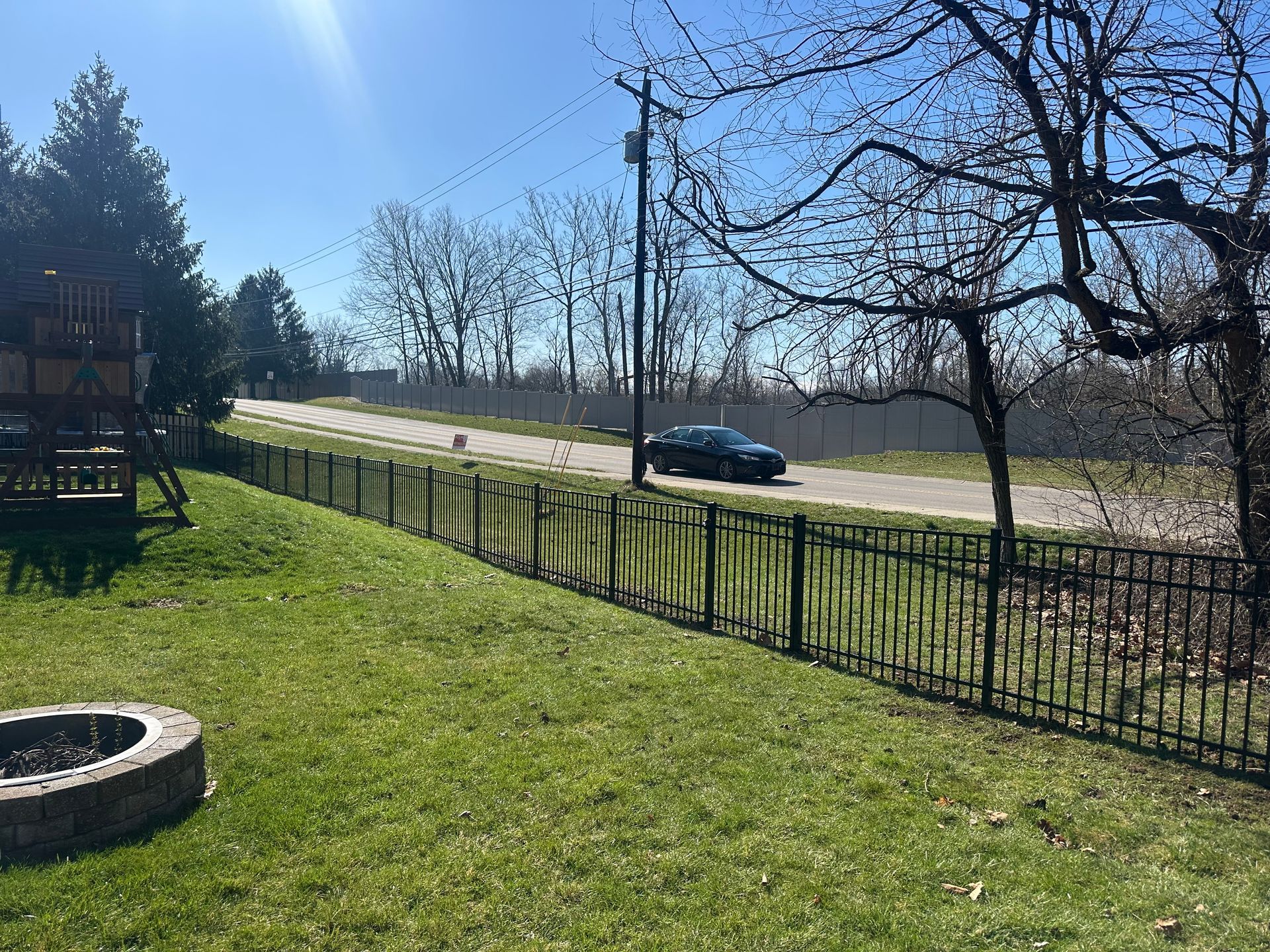 A black car drives past a black metal fence on a sunny day. Backyard with a fire pit.