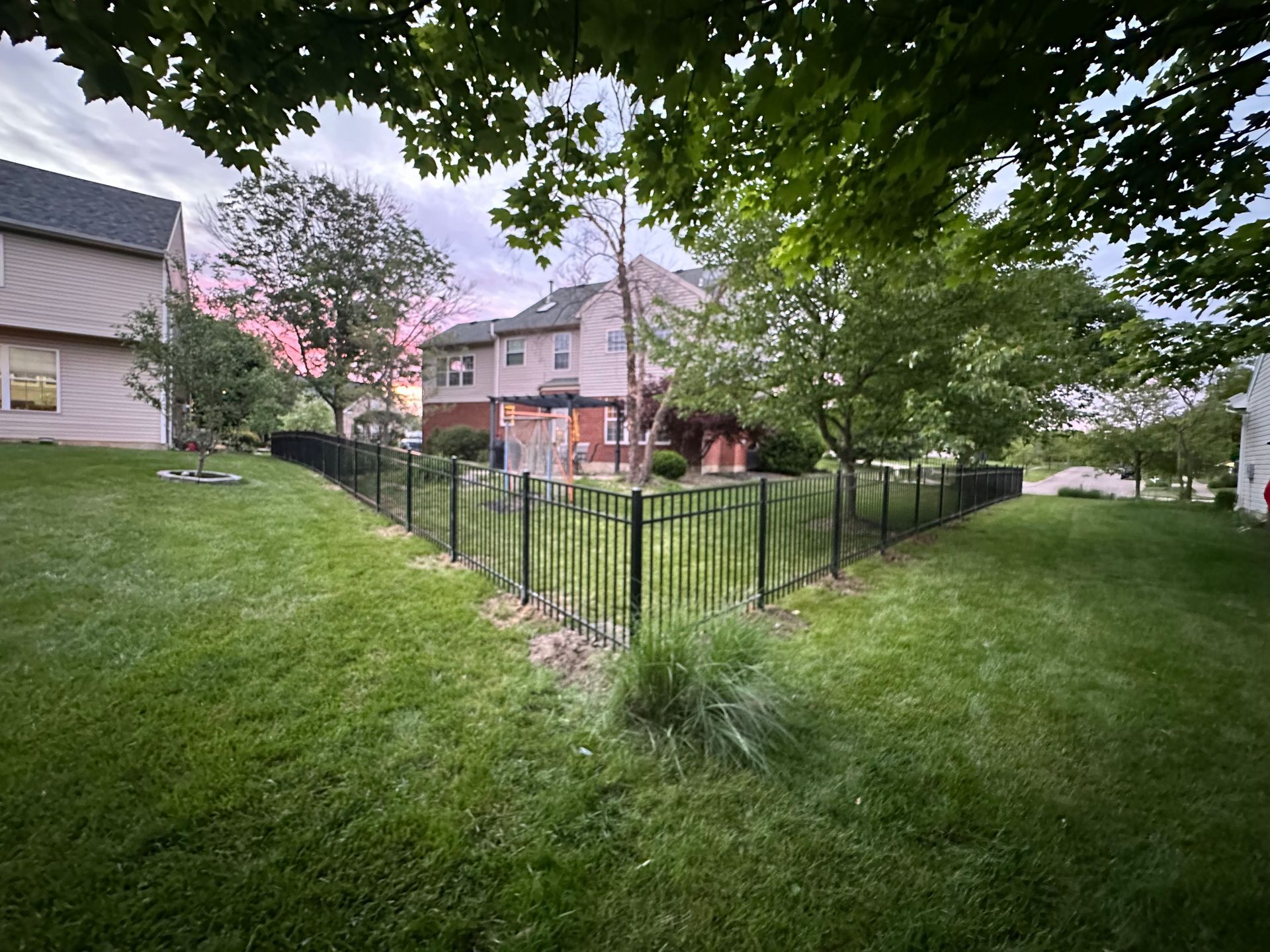 Black metal fence enclosing a small grassy area between houses with trees, under a cloudy sky.