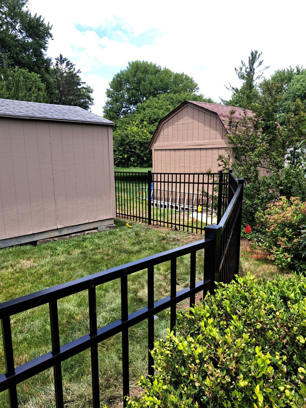 Black metal fence around a yard with two tan sheds and green trees under a cloudy sky.