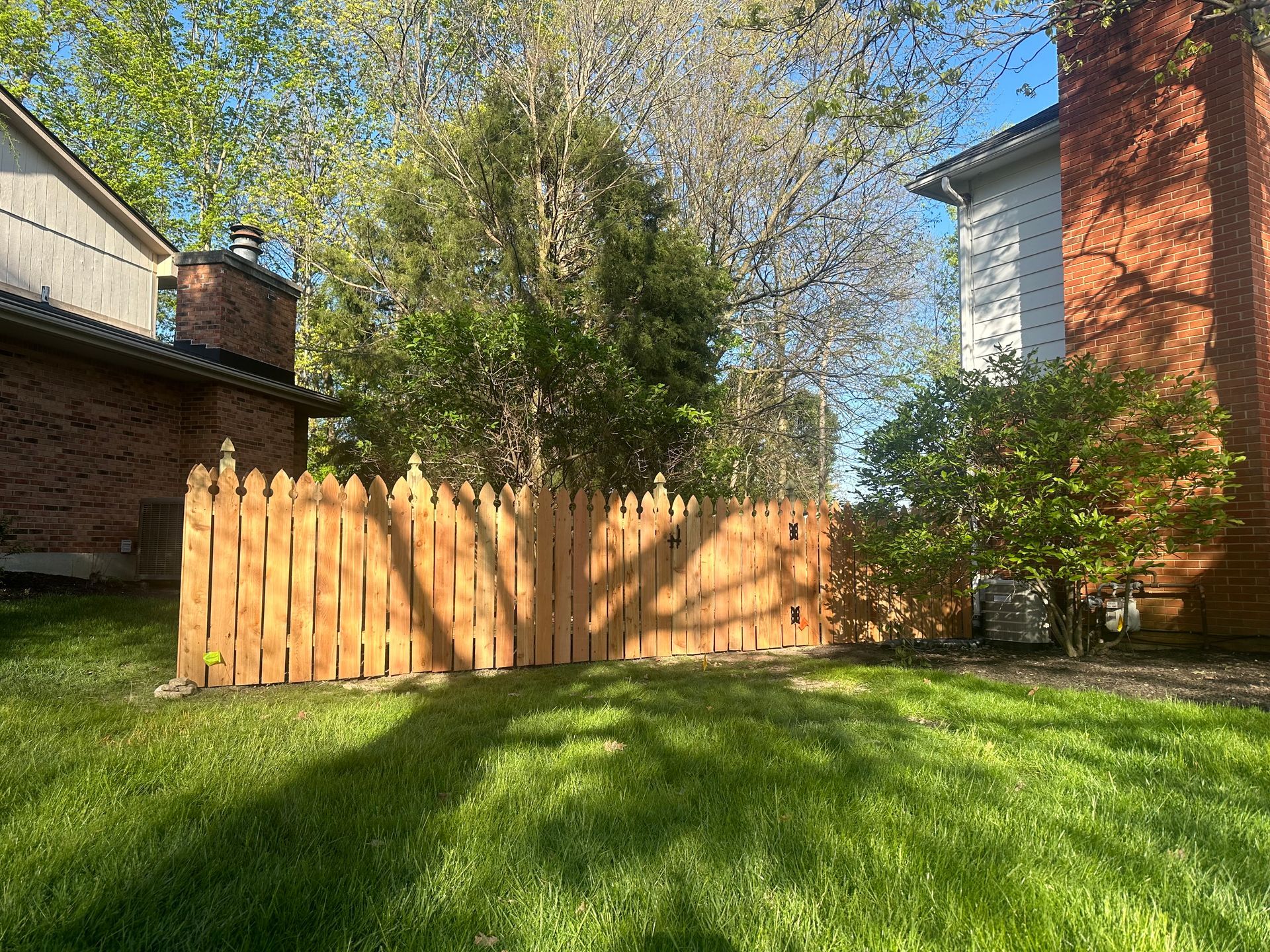Wooden fence in a backyard with green grass, trees, and parts of two brick houses in the background.