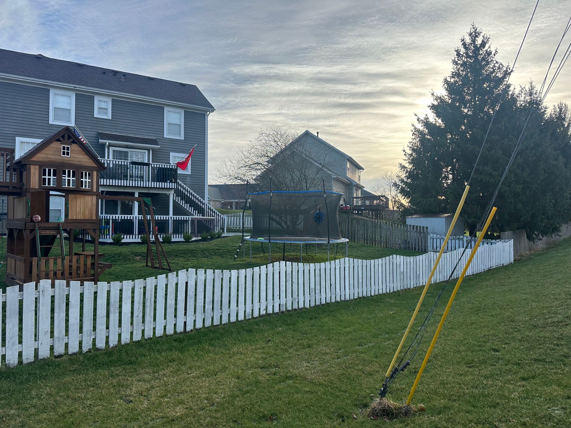 White picket fence in front of a yard with a wooden playhouse, house, and evergreen tree. Sky is overcast.
