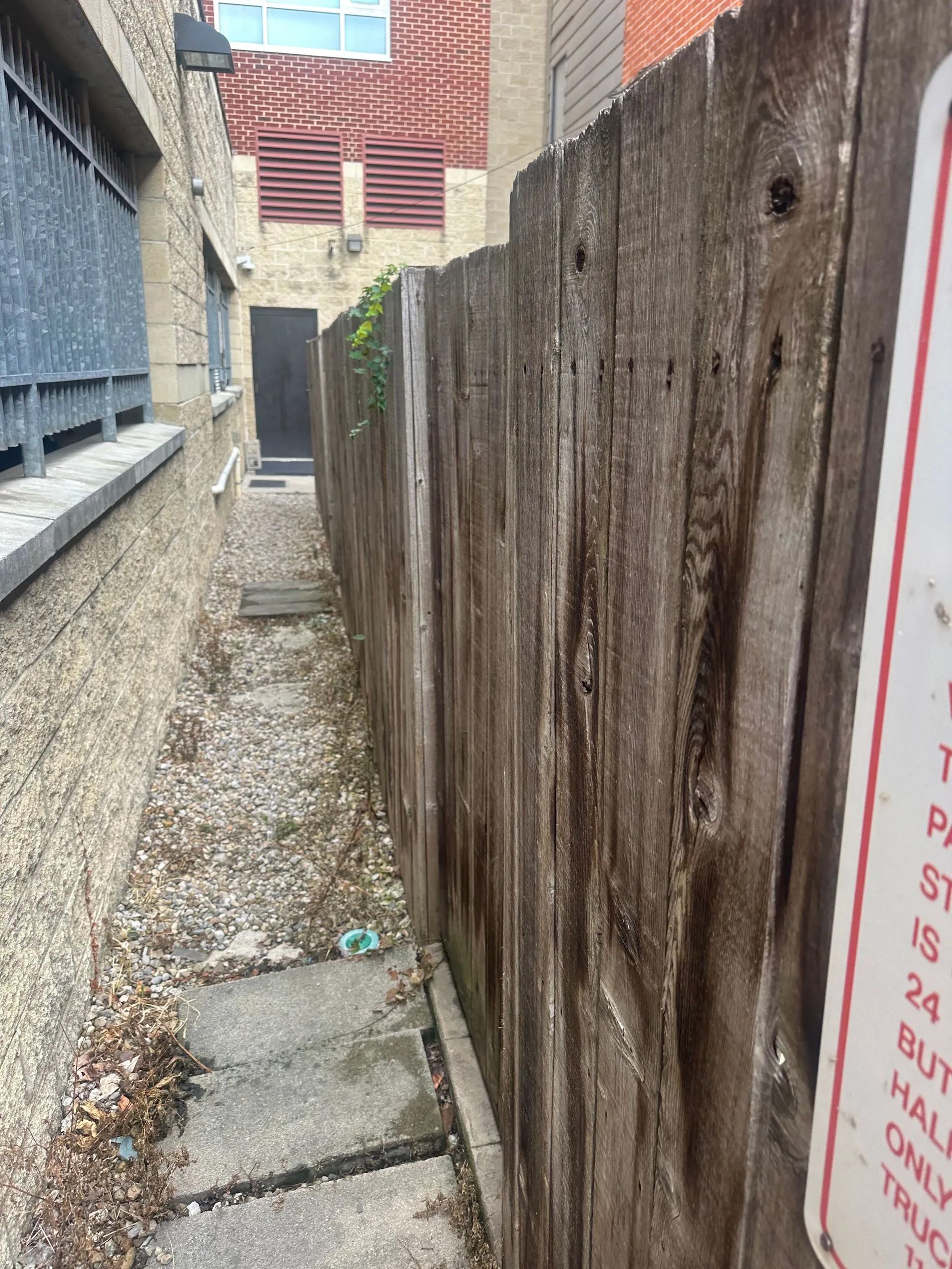 Narrow alleyway beside a weathered wooden fence and a building with a door, concrete path with gravel.