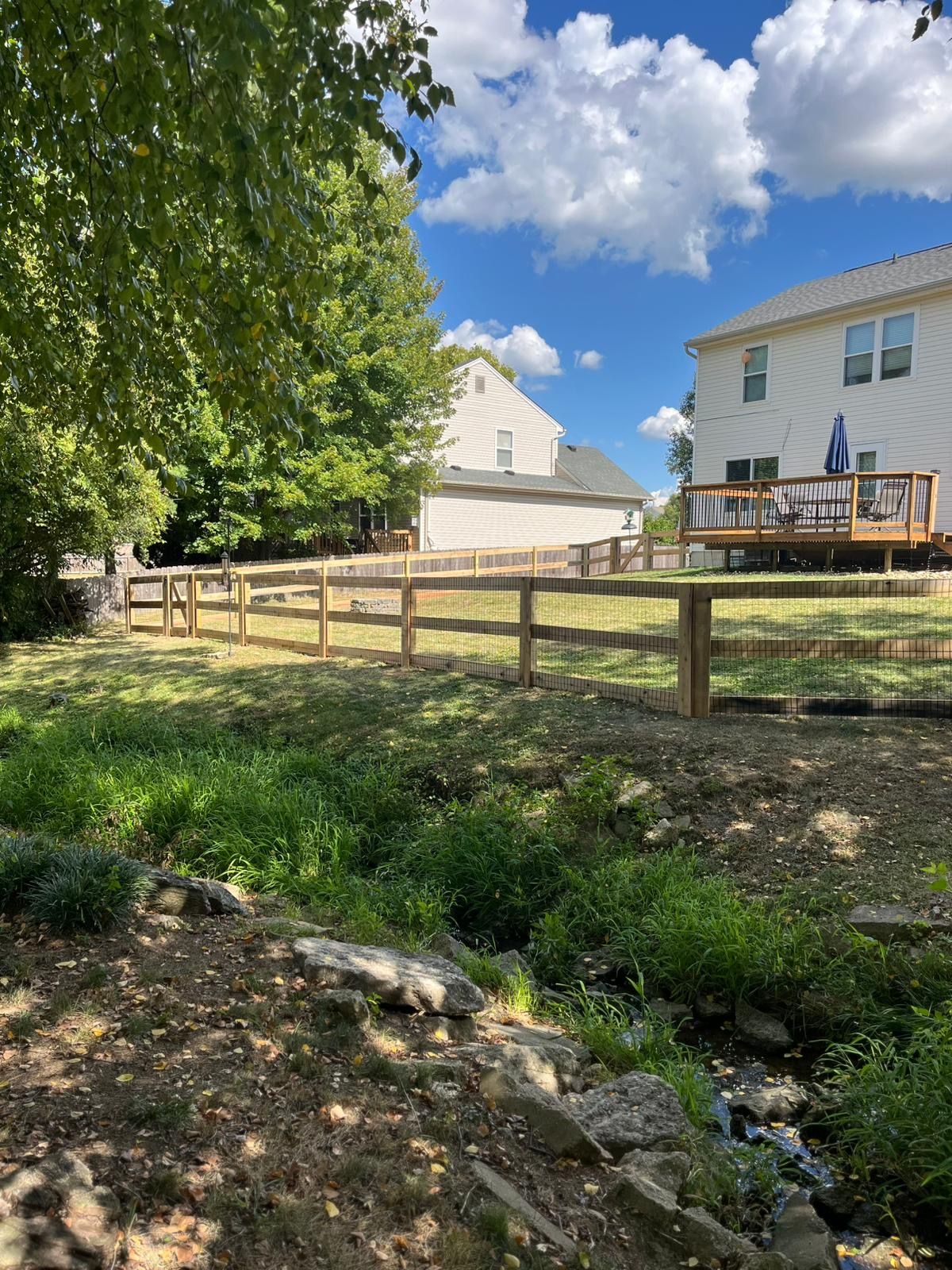 A wooden fence lines a backyard with green grass, a small stream, and two-story houses under a blue sky.