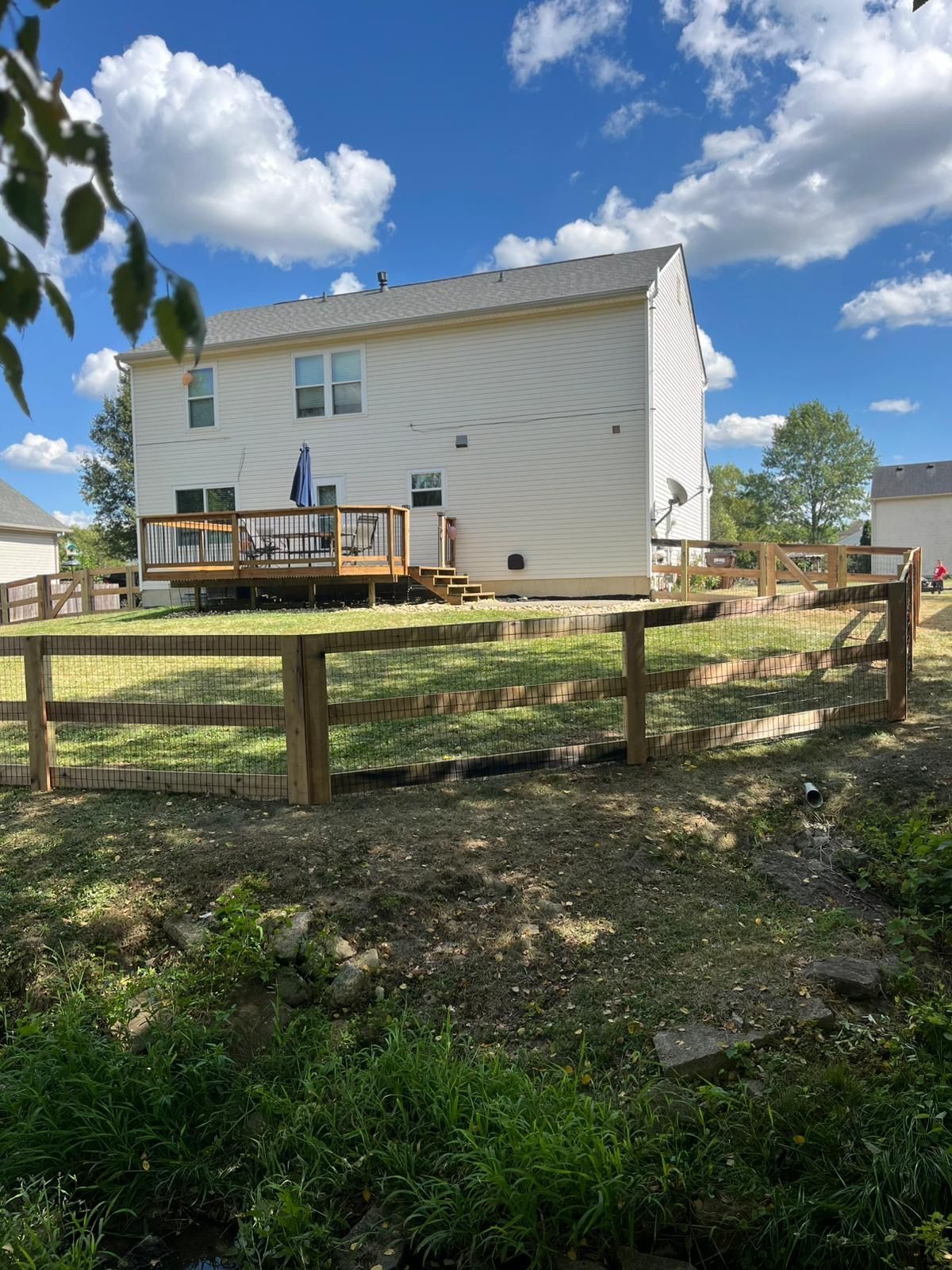 Back of a two-story beige house with wooden deck. A split-rail fence runs across a grassy yard with blue sky and clouds.