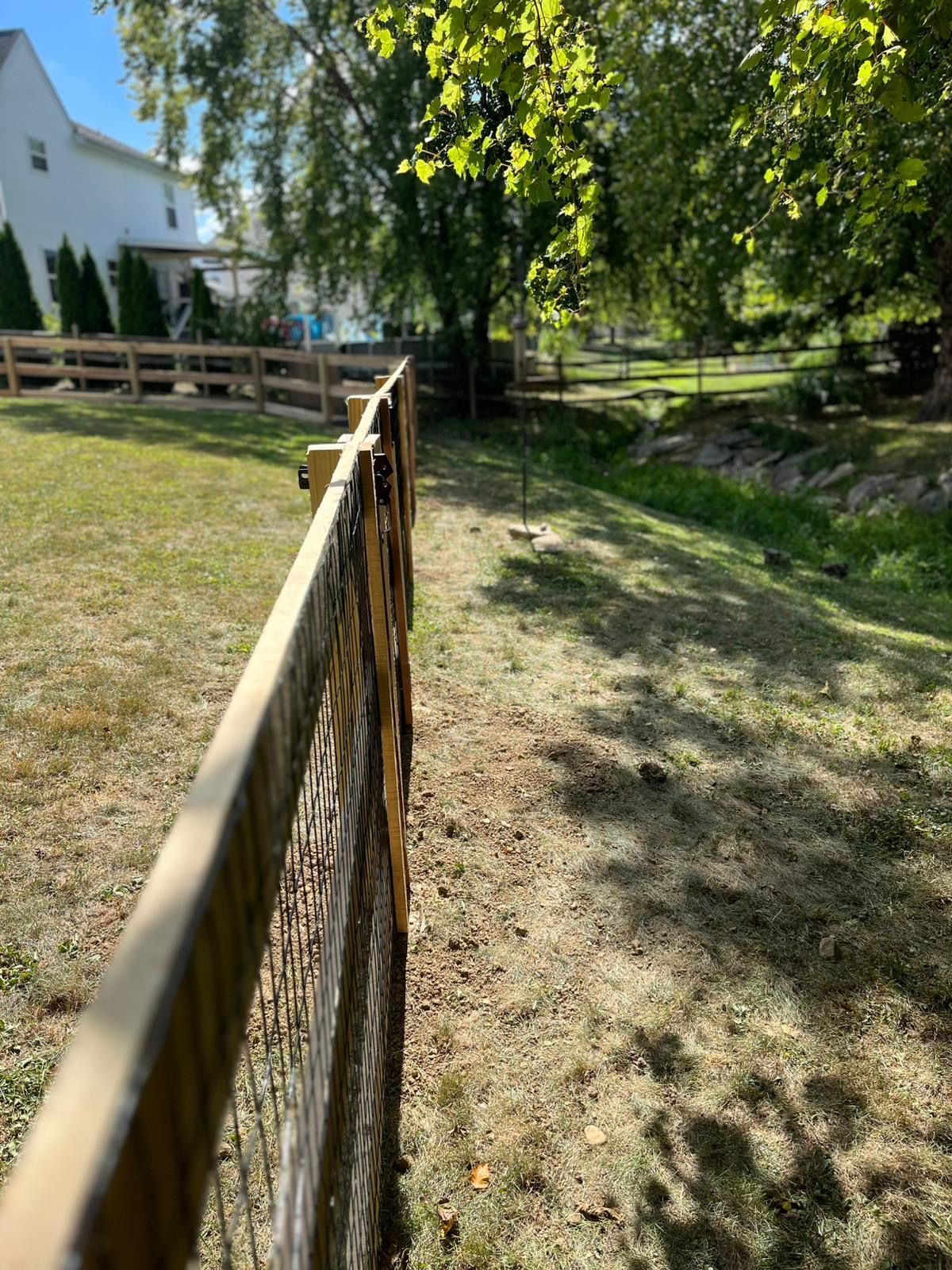 Wooden fence lines a grassy yard, beside a dry ditch with trees in the background on a sunny day.