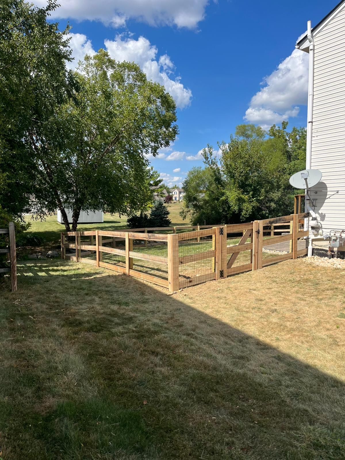 A wooden fence encloses a grassy backyard under a blue sky, with trees and a house visible.