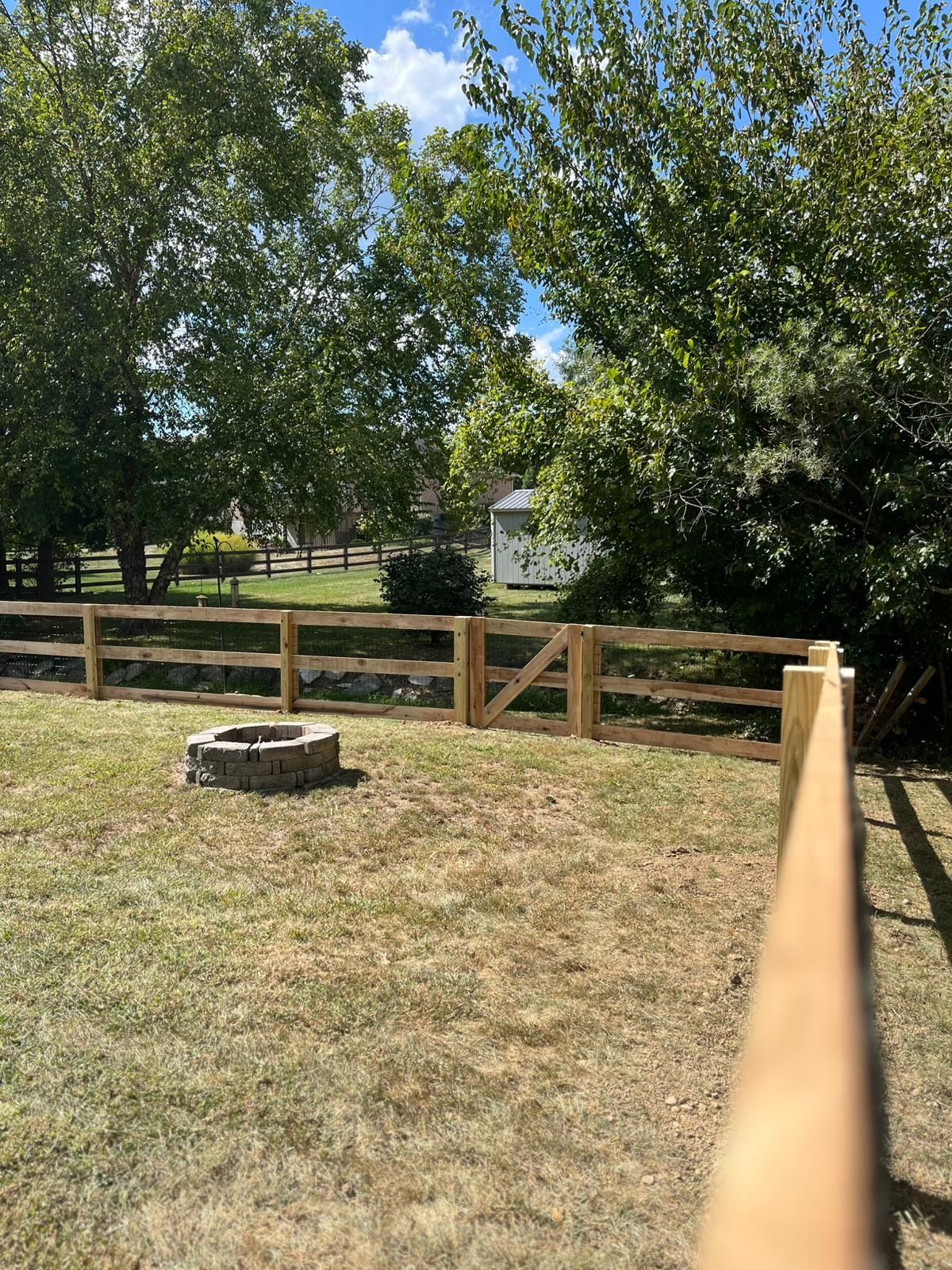 Wooden fence with a gate, fire pit in the yard, trees and house in the background. Sunny day.