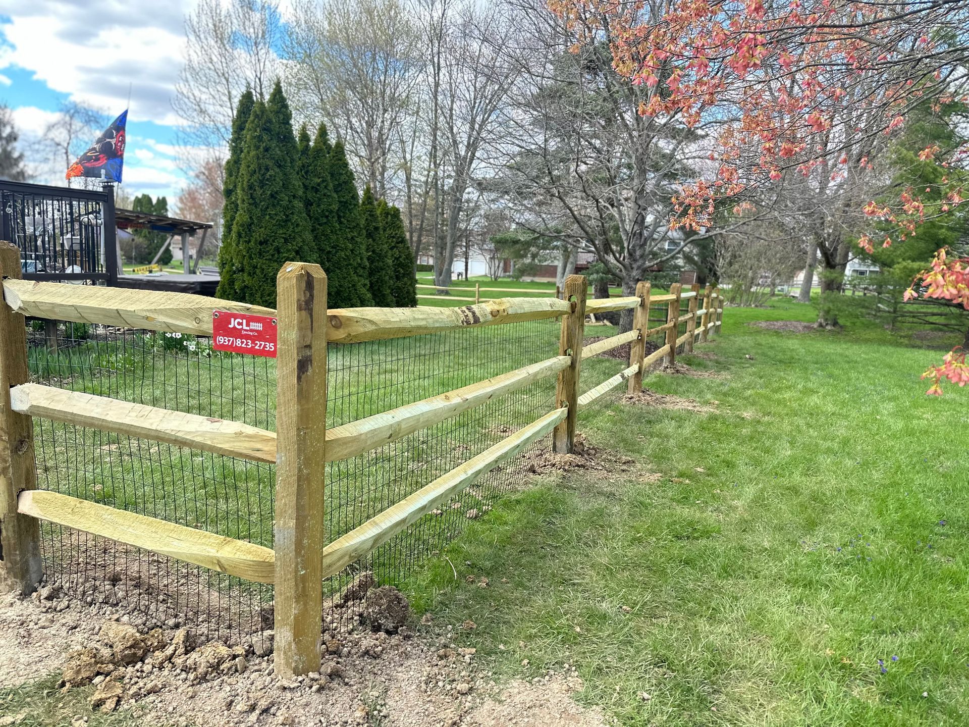 Wooden split-rail fence in a grassy yard, with a dark structure and trees in the background.