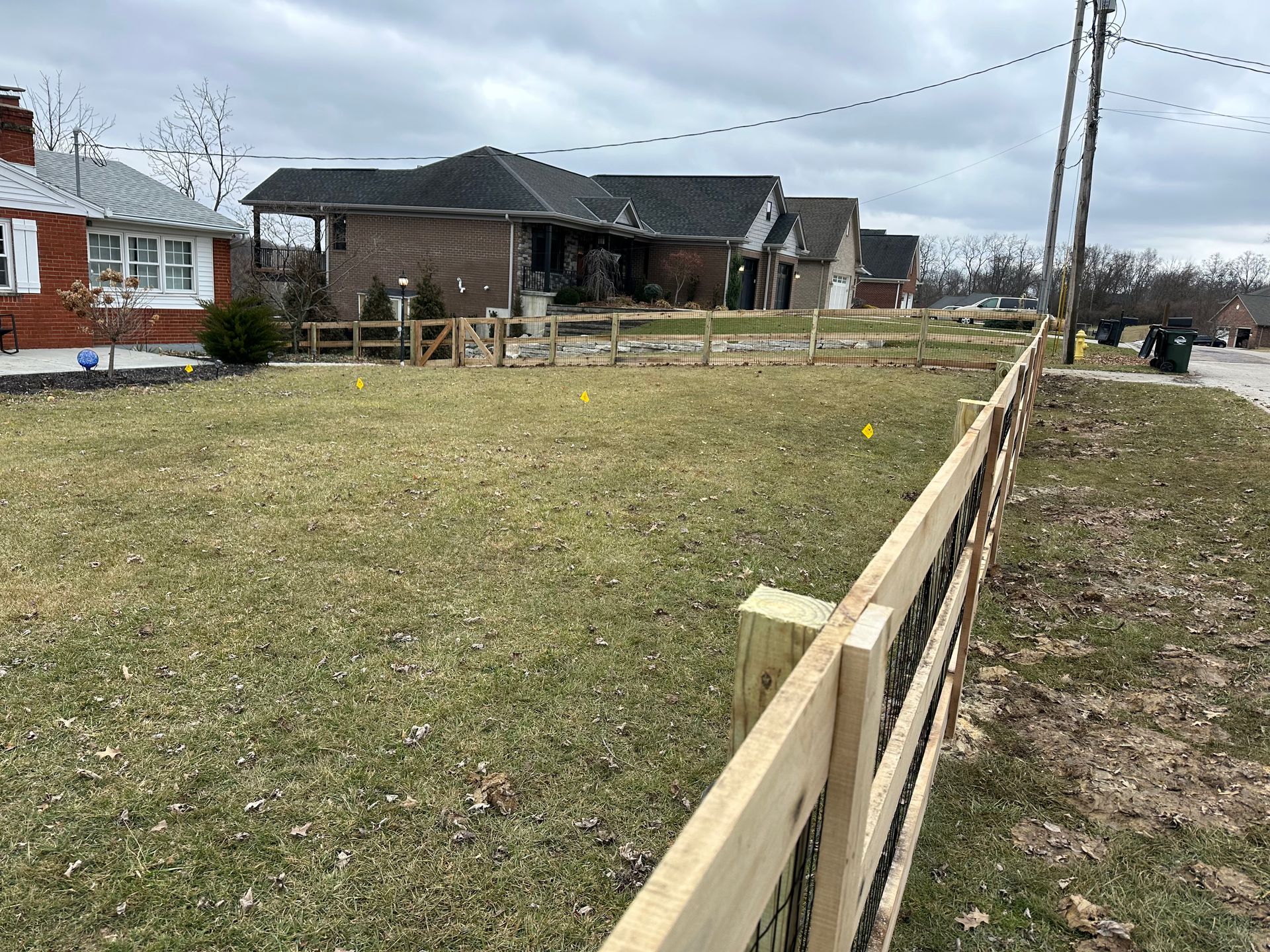 A wooden fence encloses a grassy yard in a residential area. Homes and a utility pole are in the background.
