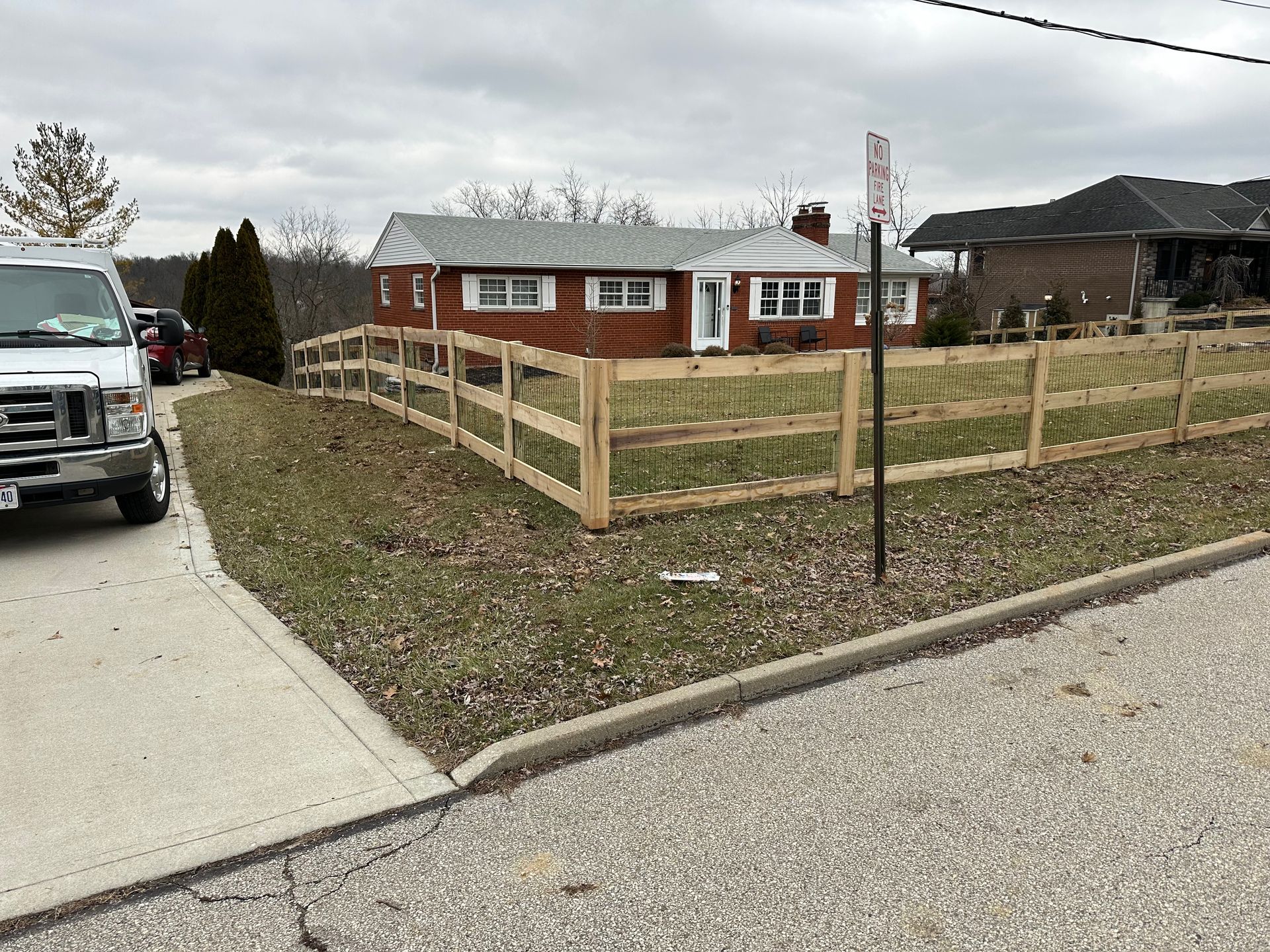 Red brick house with a new wooden fence along the front lawn on a cloudy day.