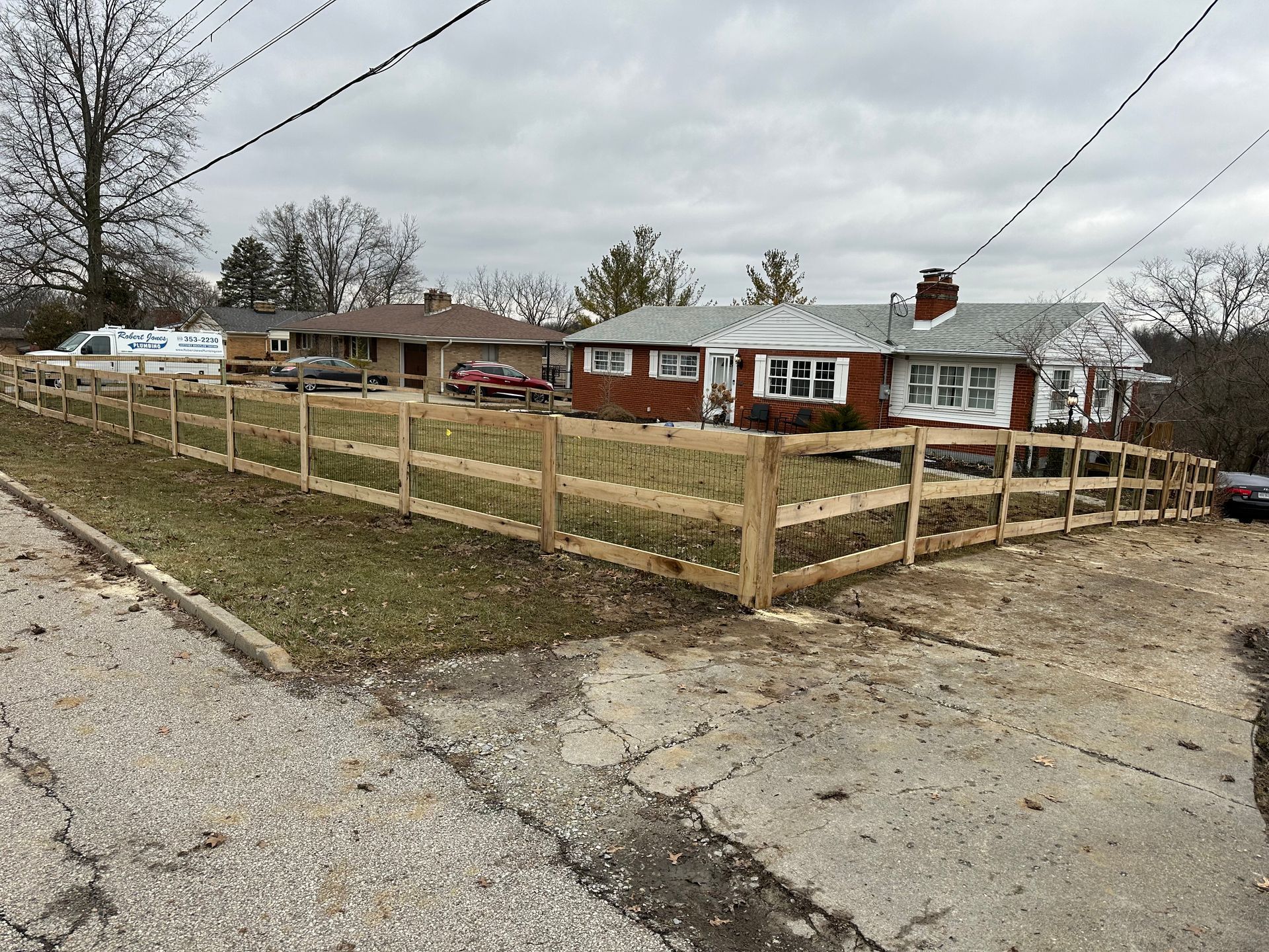A new wooden fence surrounds a residential lawn in front of a brick house on a cloudy day.