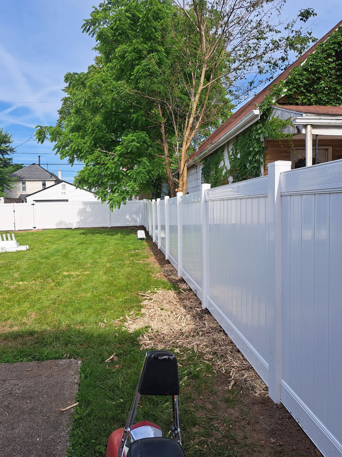 White fence in a backyard with green grass, a tree, and a building under a blue sky.