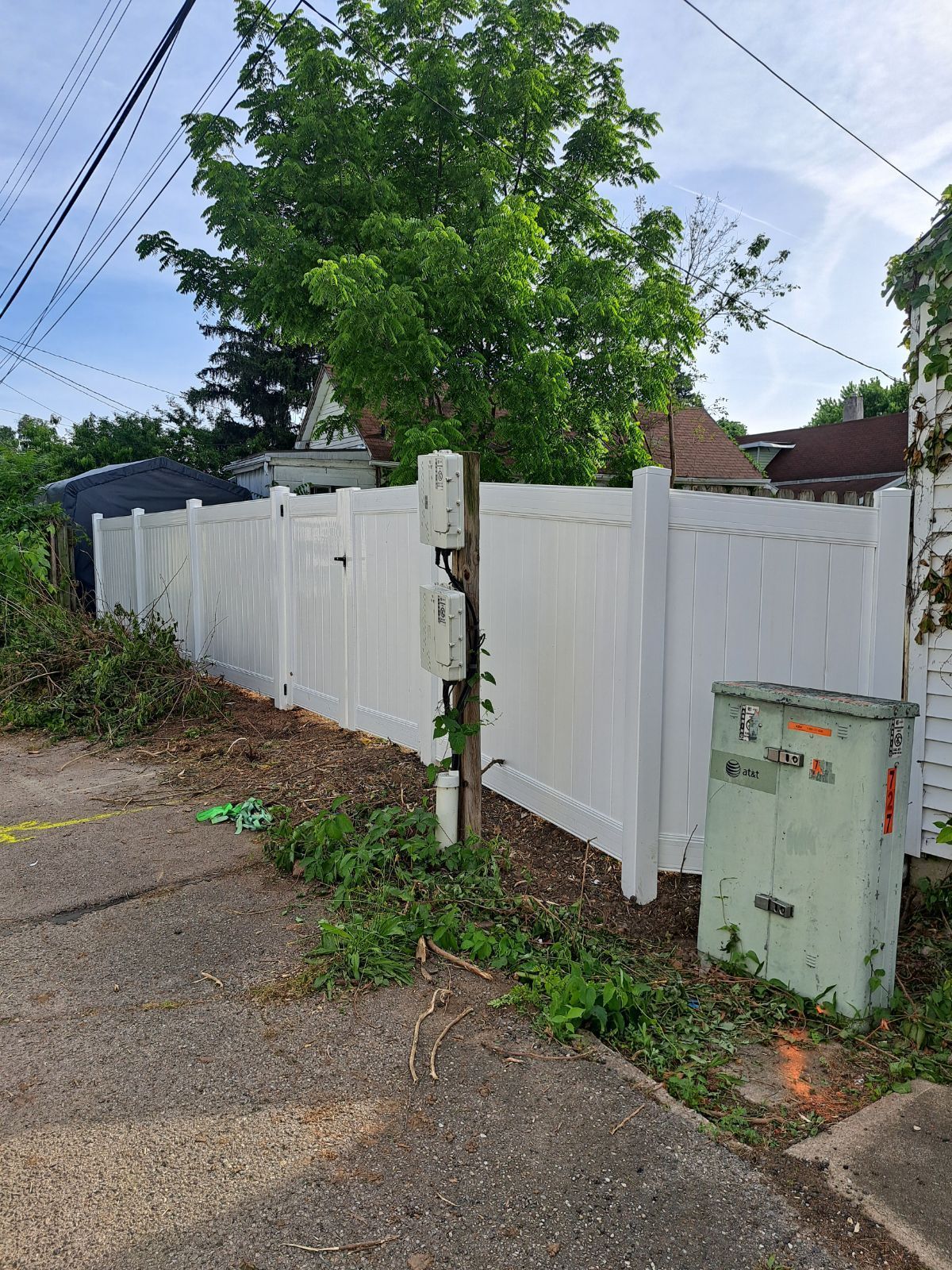 White vinyl fence along a paved area with a power box, small green plants, and a tree in the background.