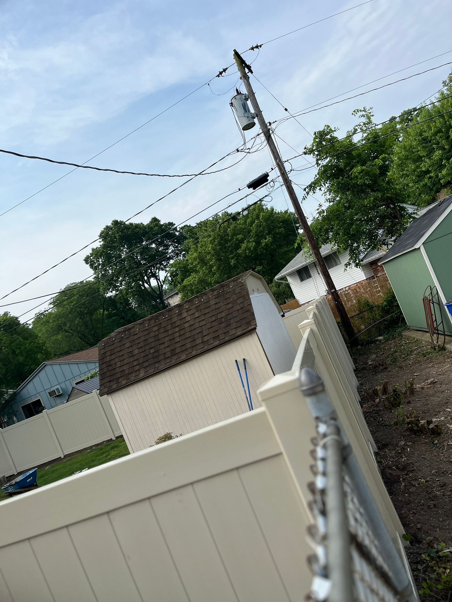 White fence in front of beige shed, power lines overhead, green trees and sky in the background.