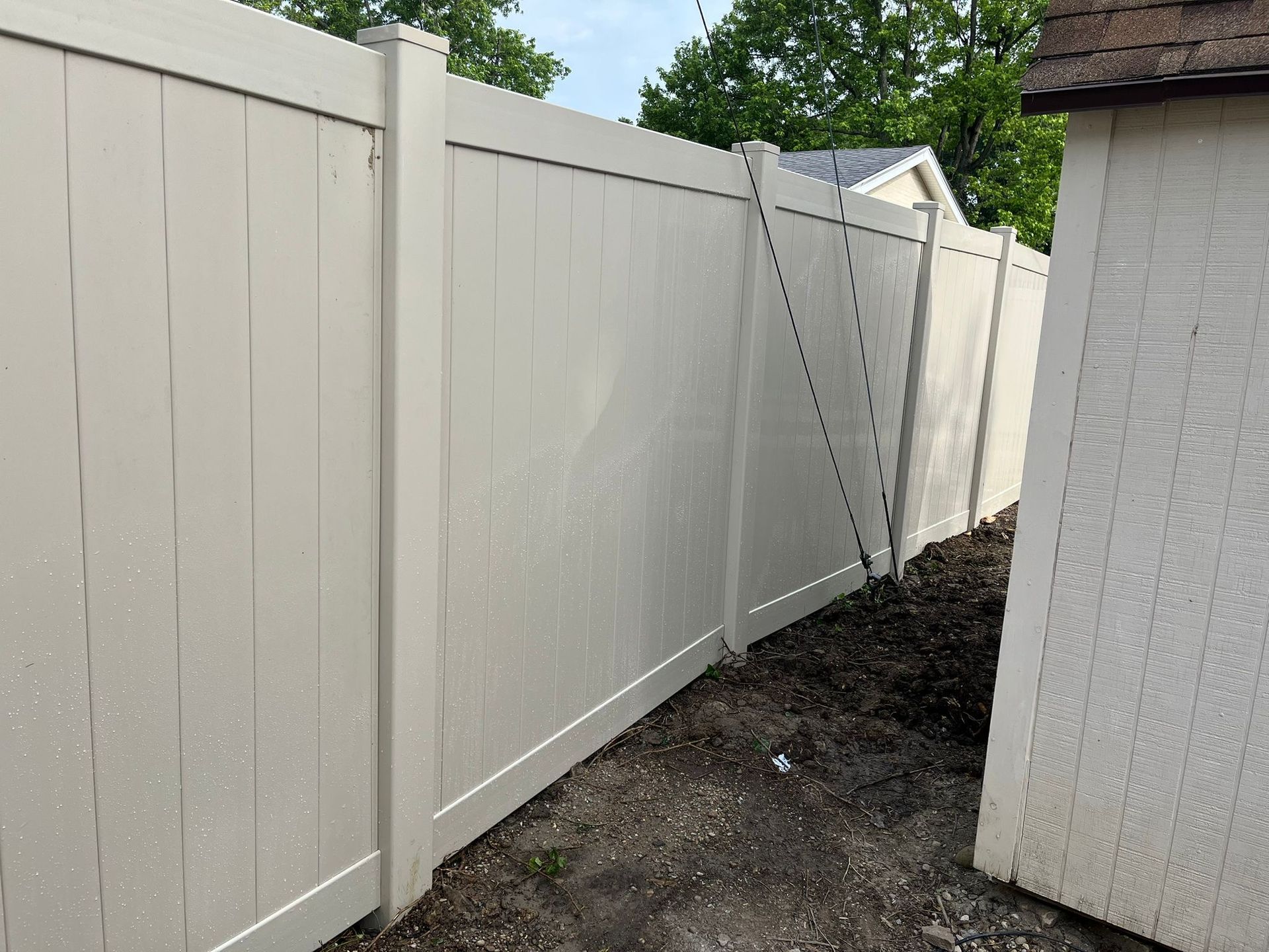 Beige vinyl fence next to a white brick building with a dark dirt patch at the base.