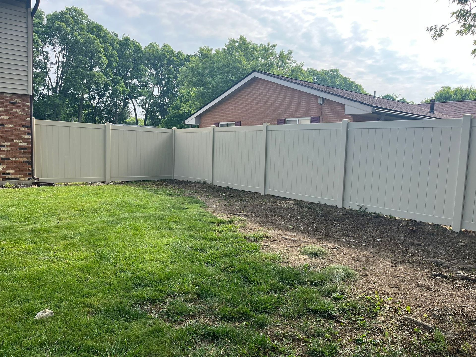 A tan vinyl fence surrounds a backyard with grass and a dirt patch. A brick house is on the left.