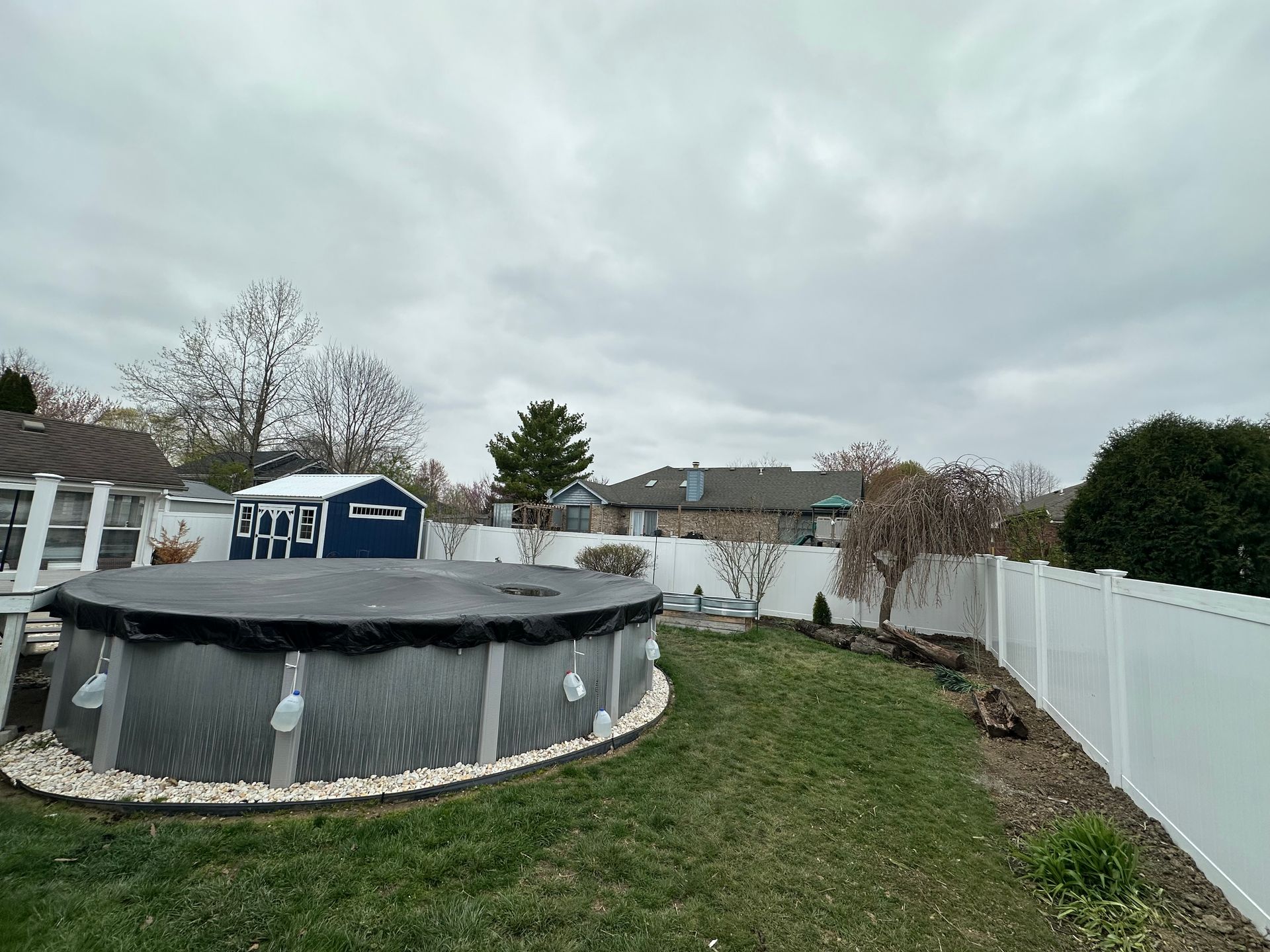 Backyard with above-ground pool covered in a black tarp, white fence, and cloudy sky.