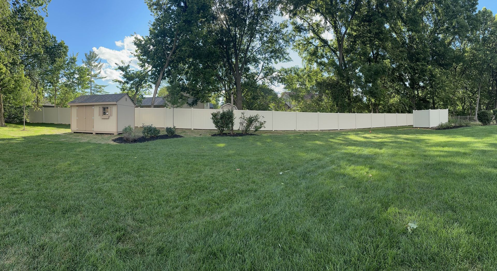 A long beige fence surrounds a grassy backyard, with a shed on the left and trees in the background.