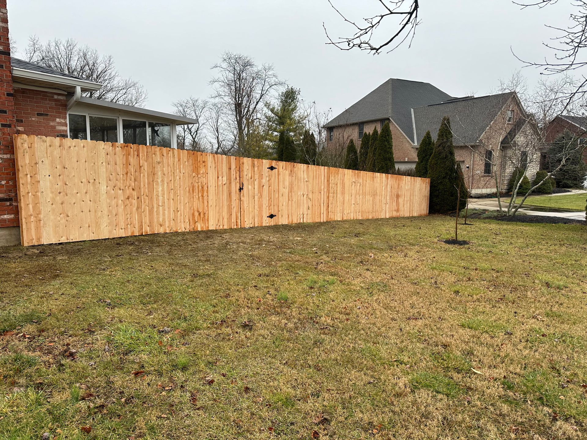 Wooden privacy fence in a backyard, beside a brick house and grassy lawn under a cloudy sky.