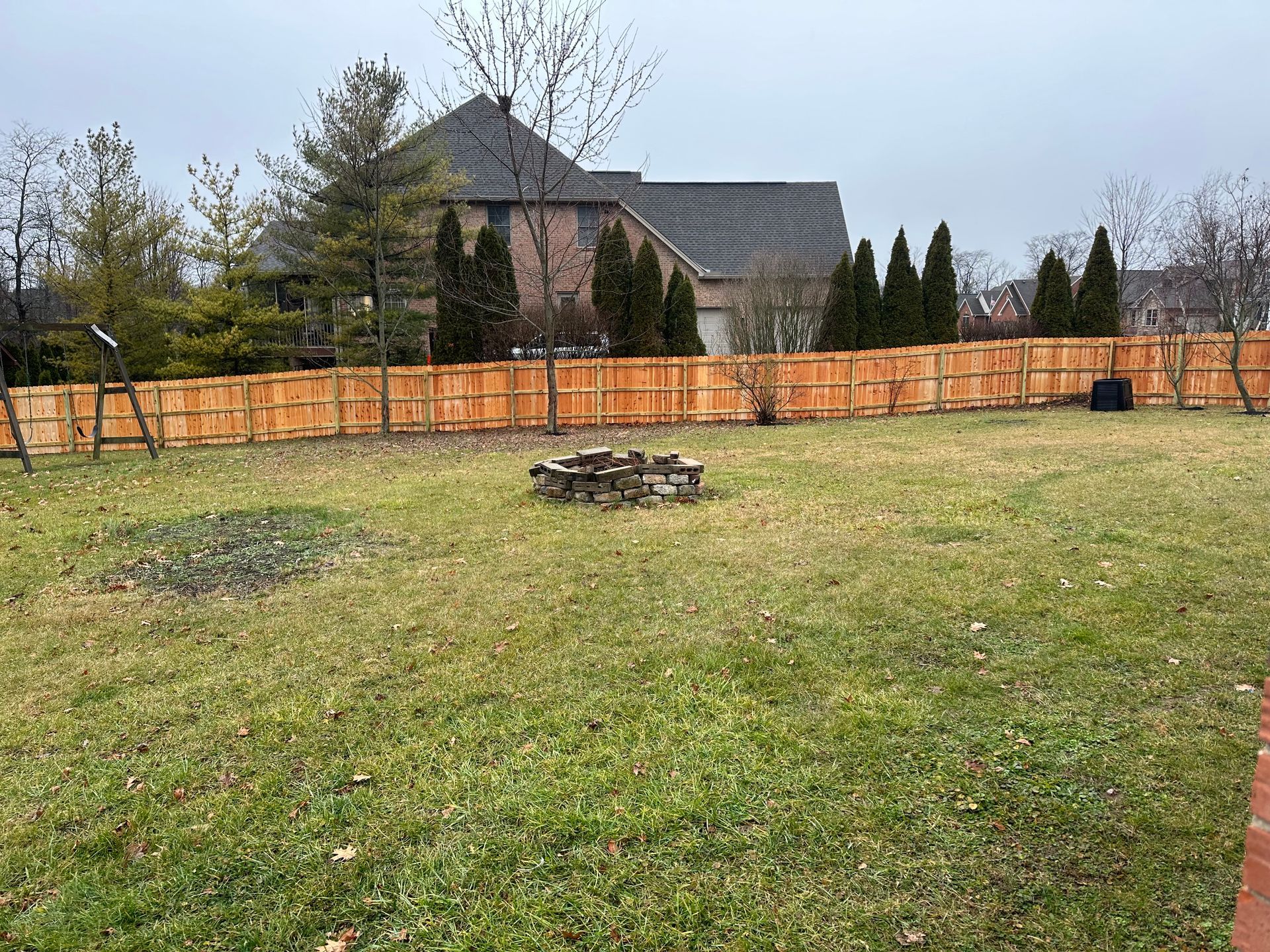 A backyard with a wooden fence, grass, and a fire pit in front of a house.