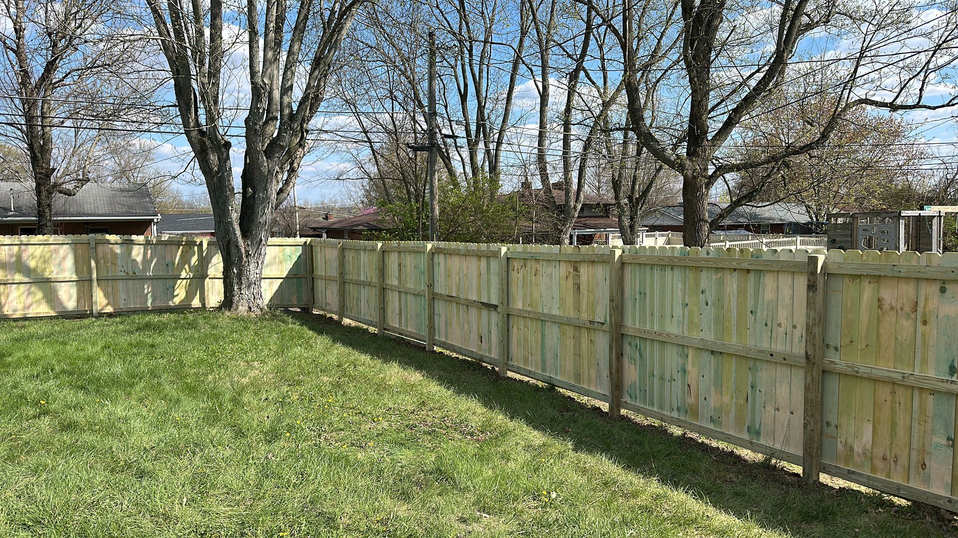 Wooden fence bordering green grass yard; trees in background; sunny day.