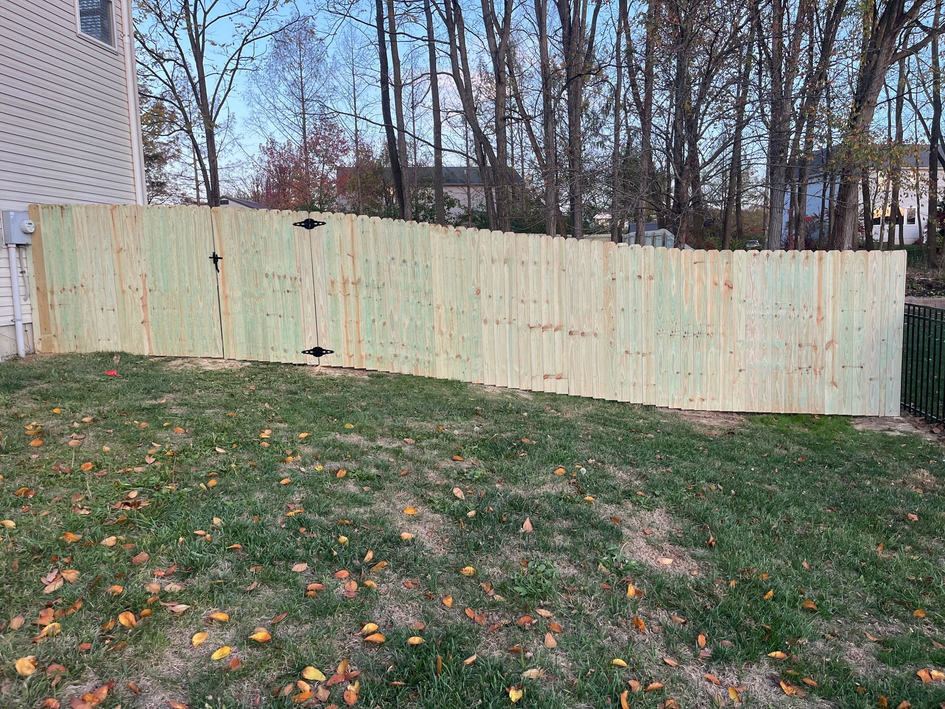 A wooden fence made of vertical boards stands on a grassy hill; trees and a house are in the background.