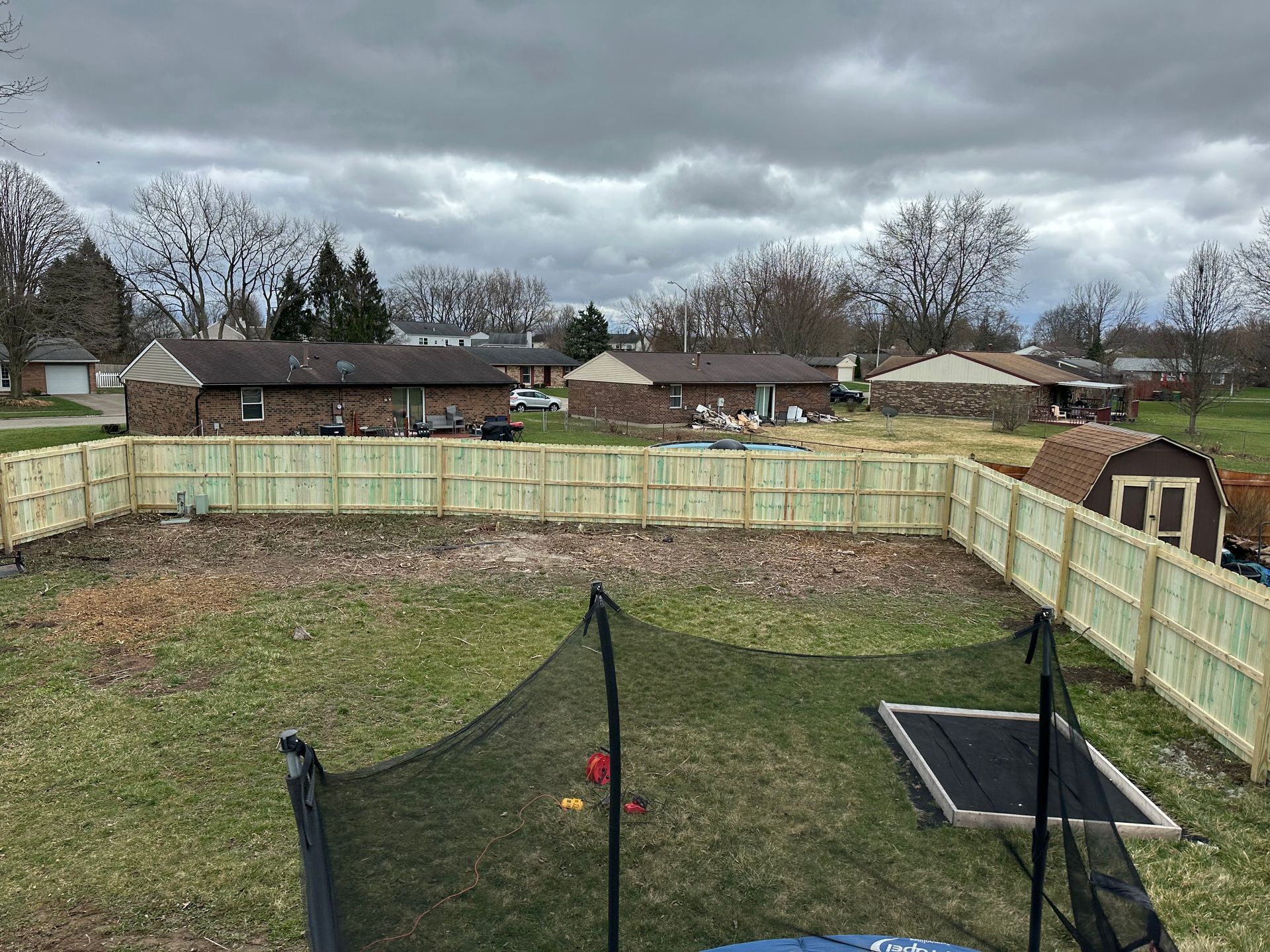 Backyard with wooden fence, trampoline, and small shed under cloudy sky.