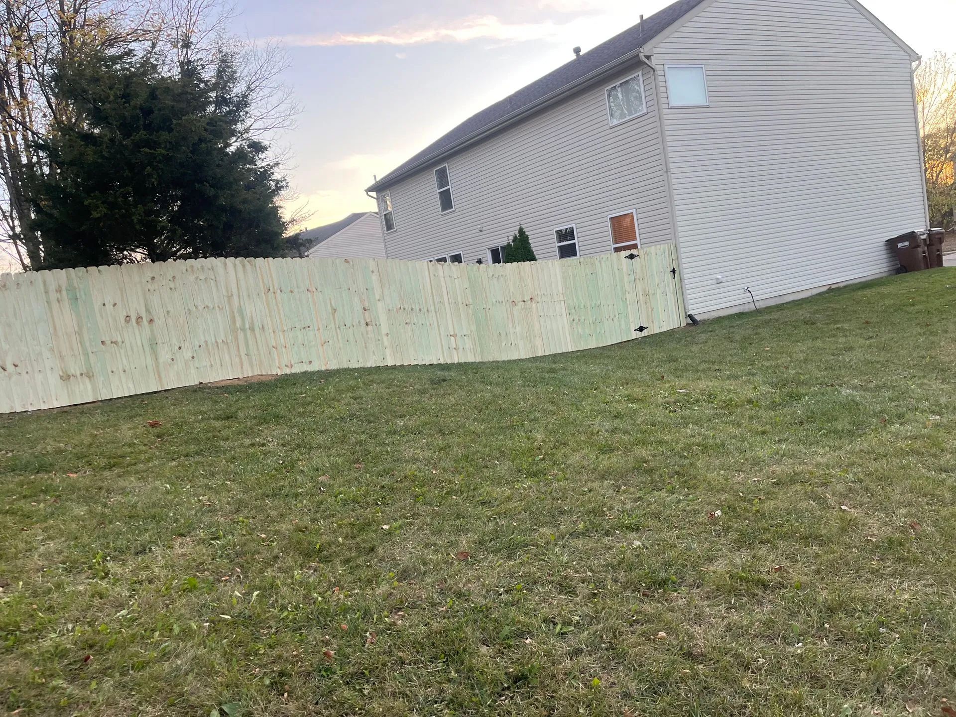 A wooden fence curves across a grassy yard, beside a two-story beige house.