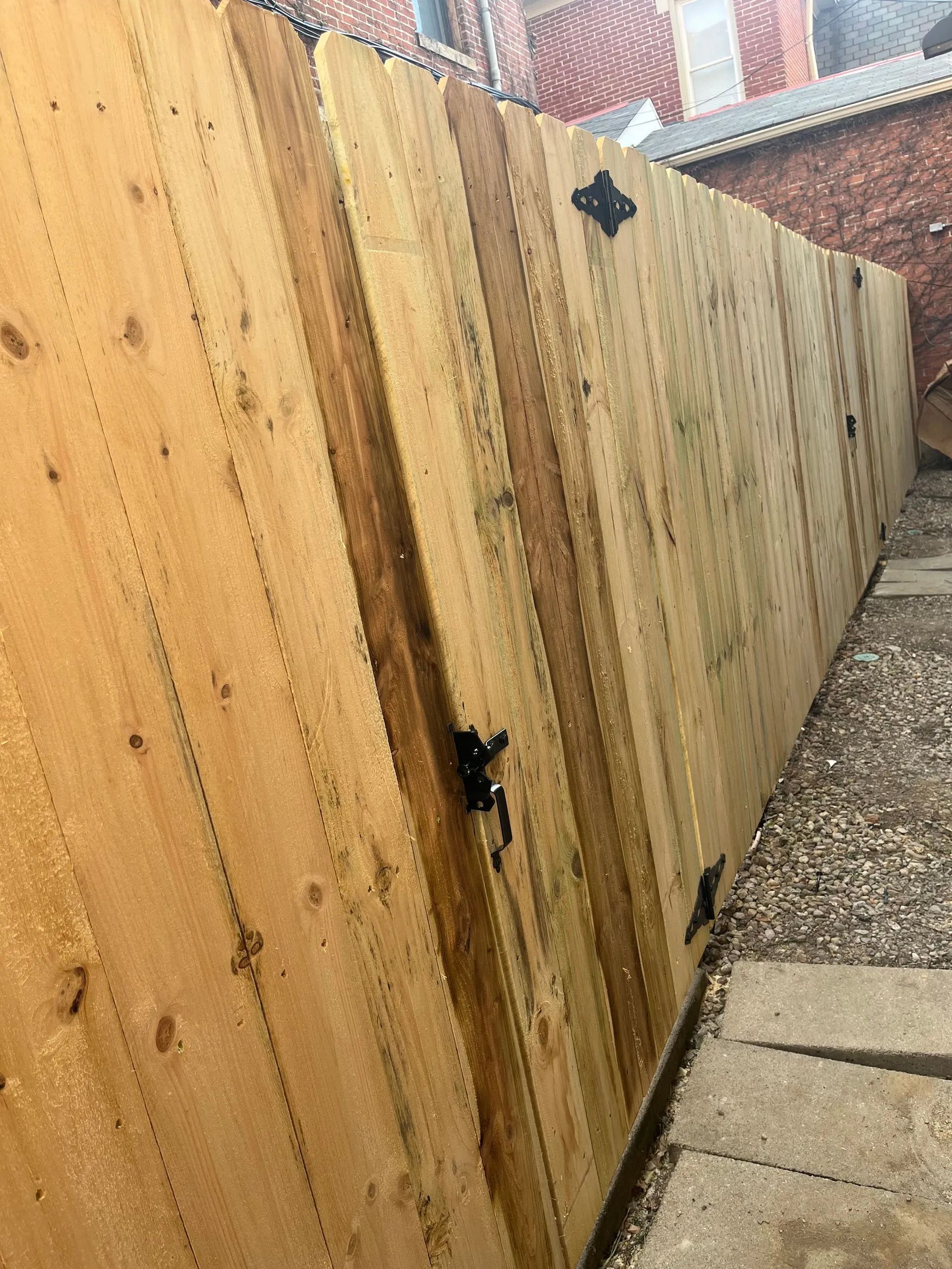 Wooden fence with black hinges, next to a gravel path and brick building.