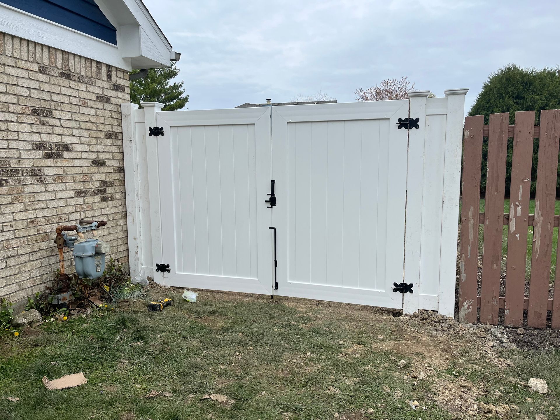 White double gate in a white fence, black hardware, next to a brick wall and brown wooden fence.