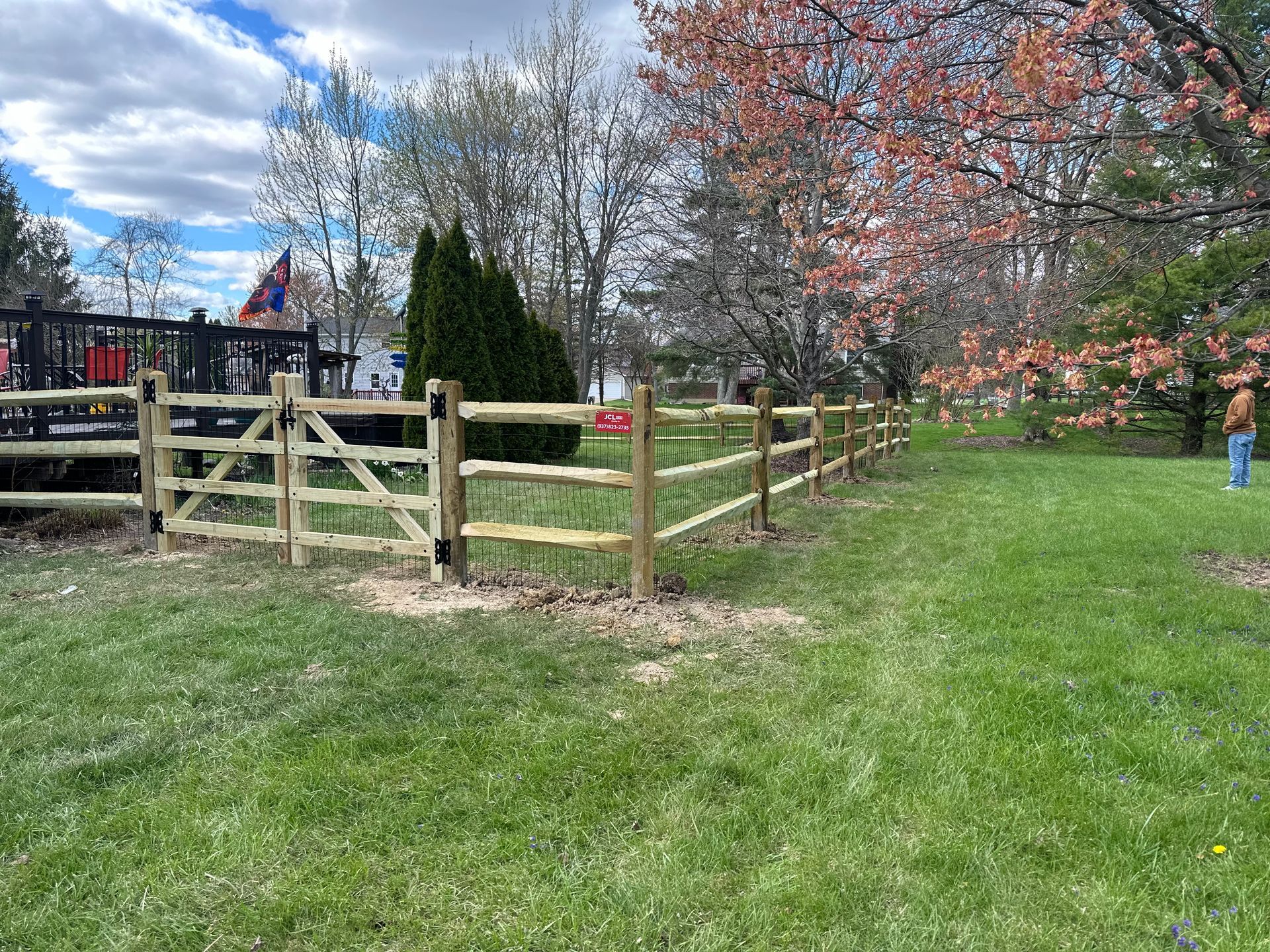 Wooden split-rail fence with gate in a grassy yard, trees in the background, blue sky.