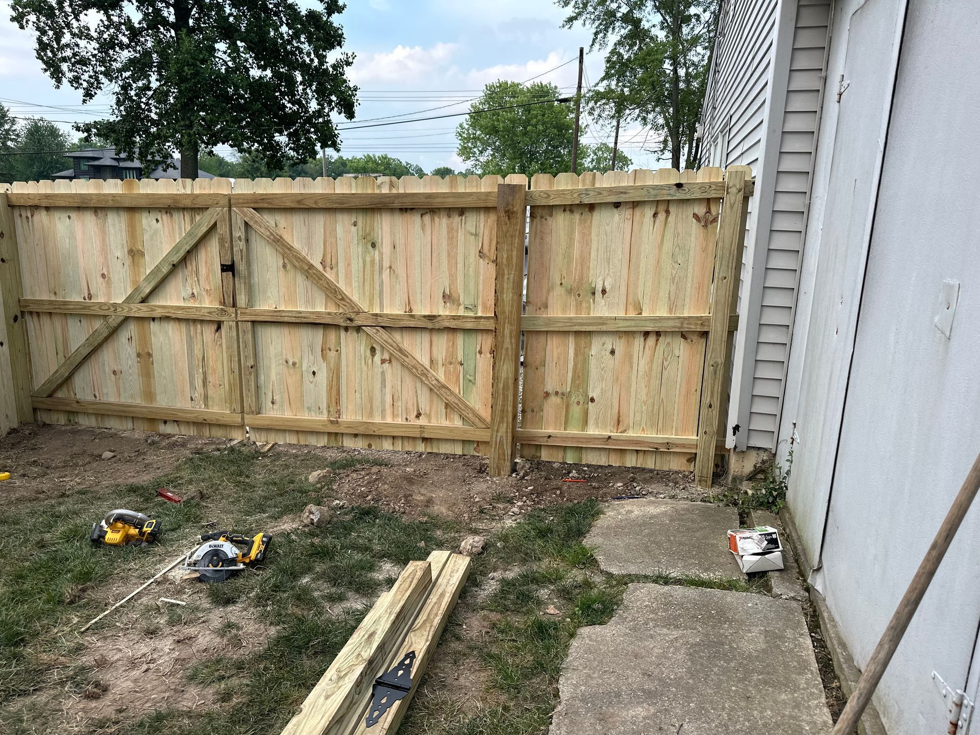 Newly built wooden fence with gate in a yard next to a building. Tools and supplies are visible on the ground.