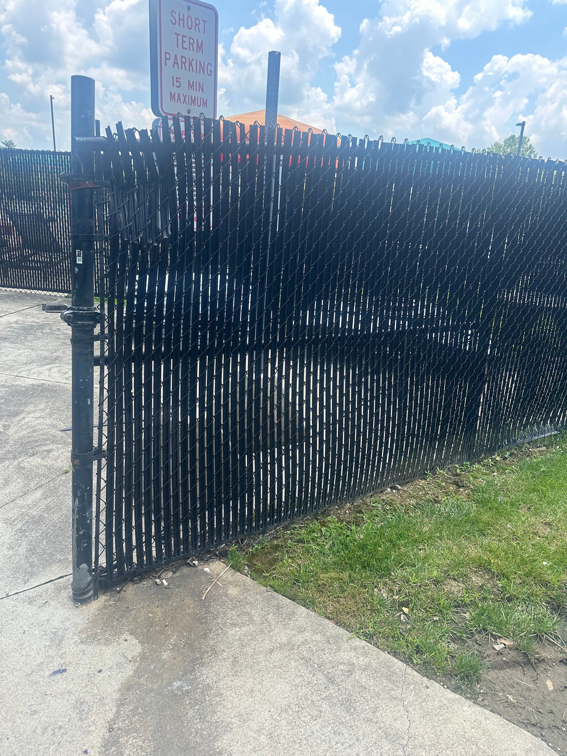 Black chain-link fence with privacy slats, gate ajar. Concrete sidewalk and green grass. Blue sky and sign in the background.