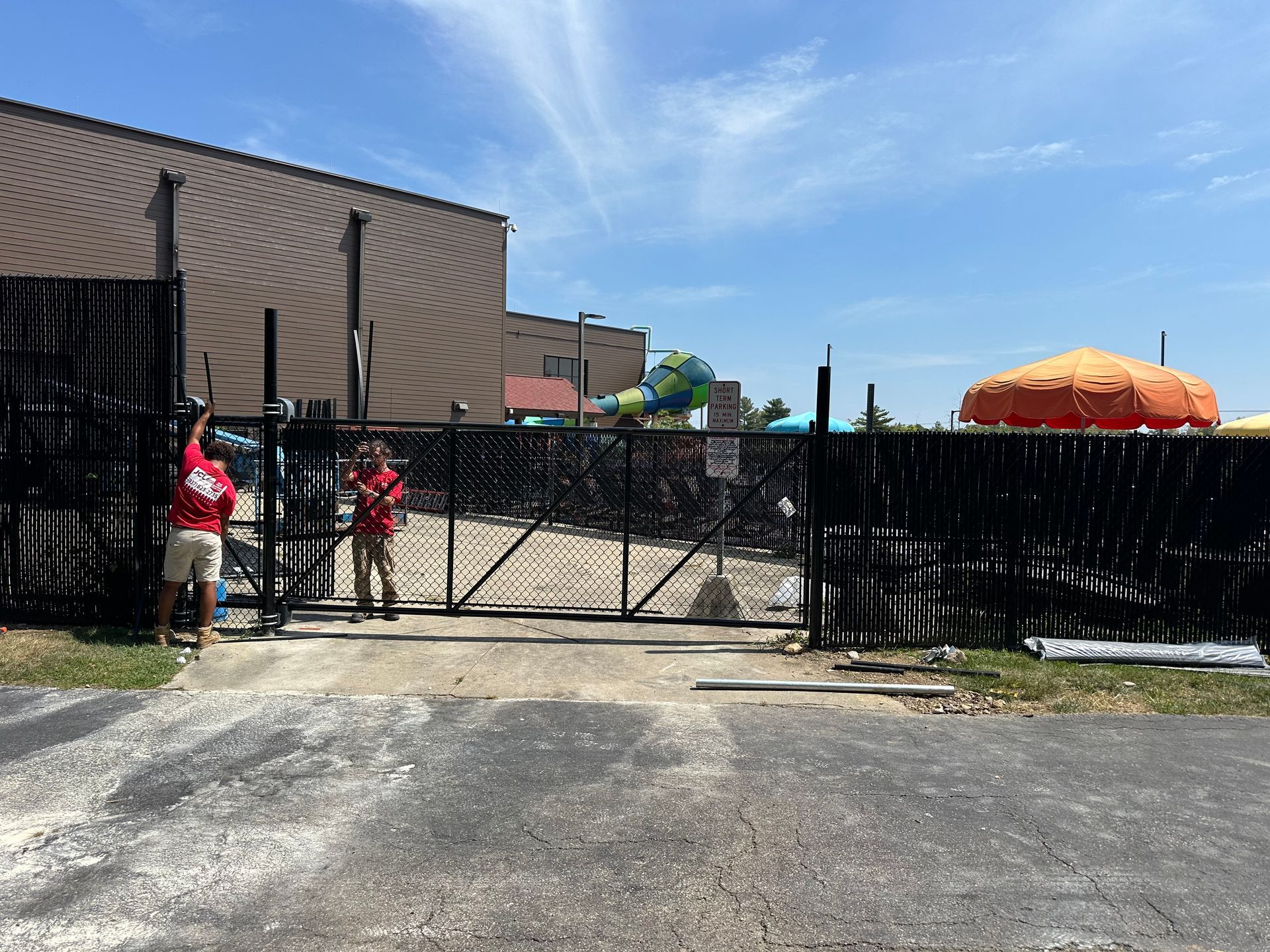 Two people opening a black metal gate to a sunny amusement park with an orange umbrella.
