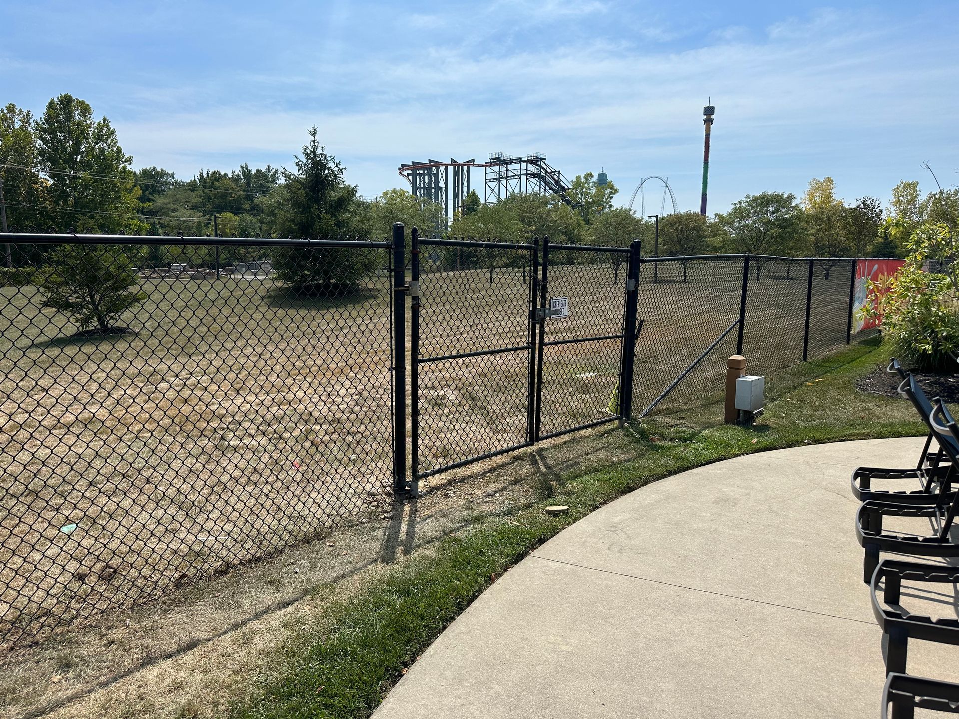 Black chain-link fence with gate, grassy area, and roller coaster in background.