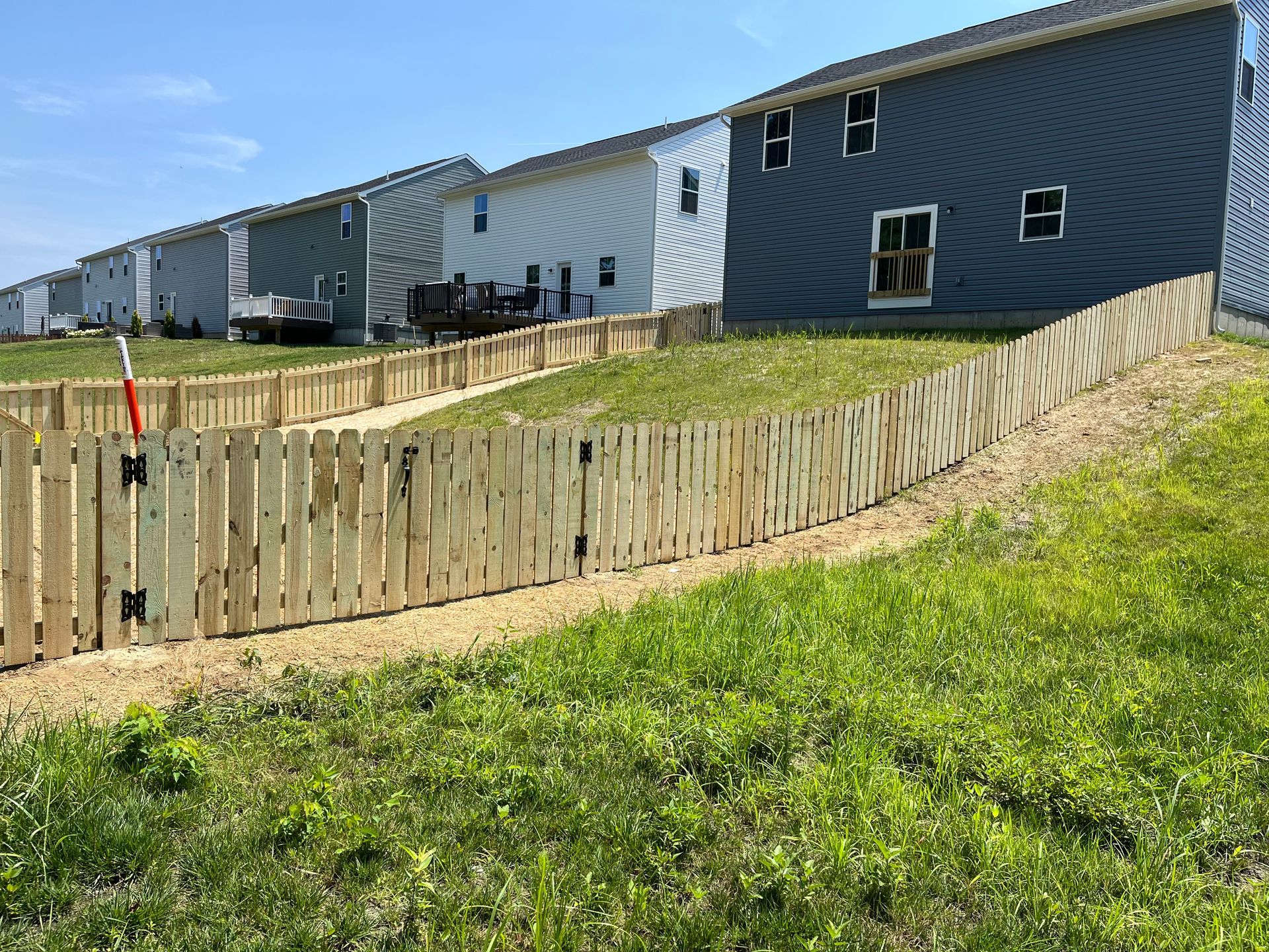 Wooden fence along a grassy slope, with houses in the background under a blue sky.